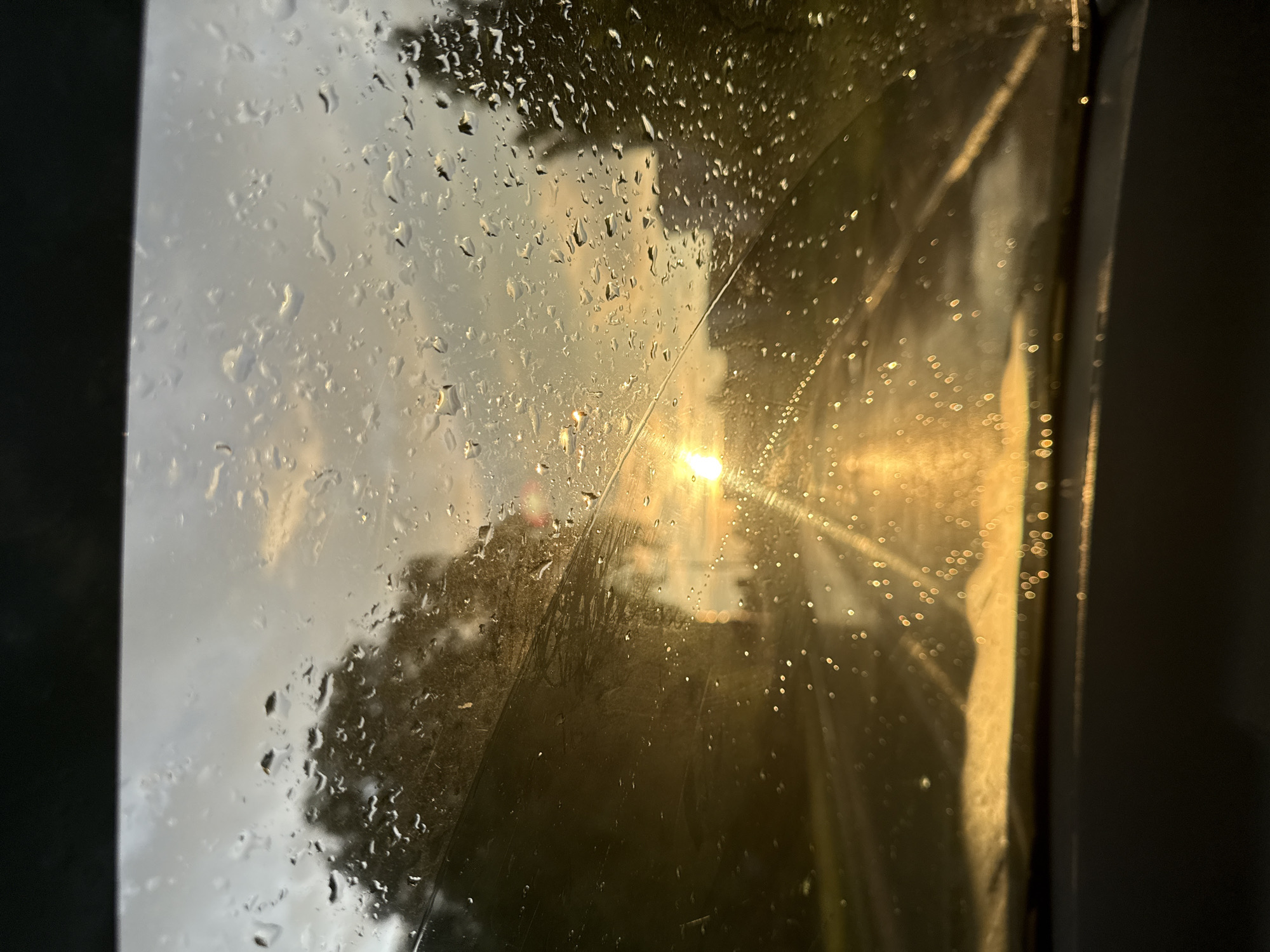 A photograph captures a view through a rain-streaked car windshield, looking out at a road during what appears to be sunset or sunrise. The sun, a bright orb, is visible in the distance, casting a golden light that reflects off the wet road and the raindrops on the glass. Silhouetted trees line the sides of the road. The bottom of the frame shows the dashboard of the car. The image does not contain any information about the city of Chaikovsky, Russia.