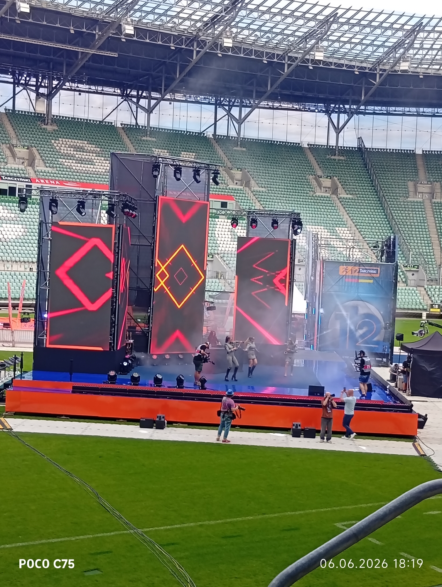 The image captures an outdoor stadium scene at Tarczyński Arena in Wrocław, Poland, likely during an event setup or performance. A large stage with an orange base and blue surface is positioned on the green grass field. The stage features three prominent vertical LED screens displaying dynamic geometric patterns in red and orange. To the right of the stage, another large screen or banner shows the "TARCZYŃSKI" logo and a large number "12". Several stage lights are mounted on the screen rigging.
On the blue stage surface, four individuals, potentially performers, are visible on the left side, dressed in light-colored outfits and dark boots, with two holding prop objects. Smoke effects are emanating from the stage. On the right side of the stage, a person wearing a dark vest and shorts operates a camera mounted on a stabilization rig. Off-stage, on the grass, two individuals are standing and holding up smartphones or cameras towards the stage. Another person, dressed in blue, stands further left on the grass, holding a prop similar to those on stage. Electrical cables are stretched across the green grass in the foreground. In the background, green and white stadium seating tiers are visible, largely unoccupied, under a large metal roof structure. A black tent and additional personnel are located near the large "12" screen. The image is watermarked with "POCO C75" and a timestamp of "06.06.2026 18:19".