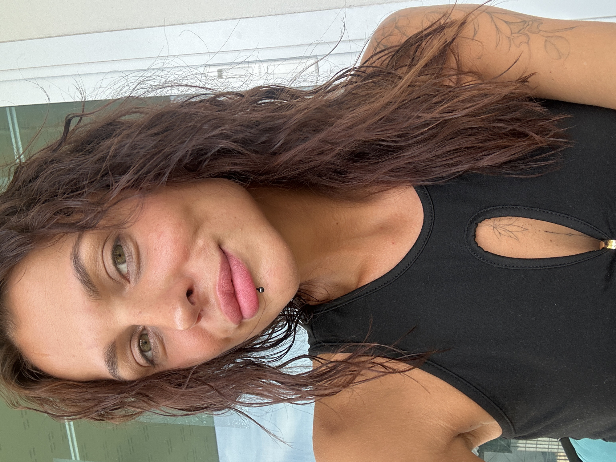 A woman with medium-length, wavy brown hair and green eyes is pictured in a close-up, selfie-style shot. She is wearing a black, sleeveless top with a cutout in the décolletage area, revealing a tattoo on her chest. She has a lip piercing just below her lower lip and is wearing light pink lipstick. The background appears to be an outdoor setting with vertical white elements that could be part of a building or railing, and a blurred background of a window with the sky visible through it. The lighting suggests it is daytime, possibly overcast. The image does not contain any information about the city of Ko Samui District, Thailand.