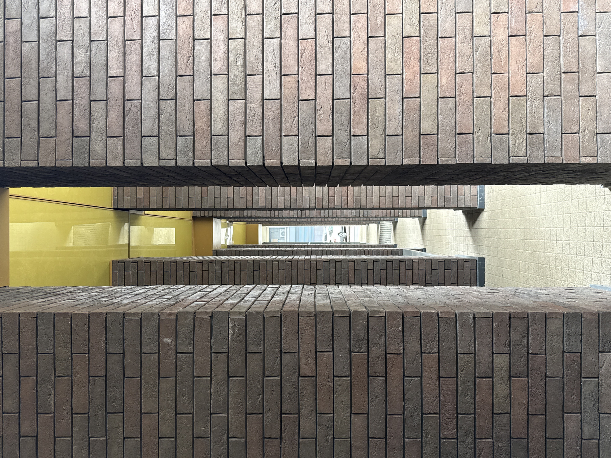 The image captures a unique architectural perspective within a building or complex in Moscow, Russia. The scene is devoid of people and focuses entirely on the structure itself, creating a strong sense of depth and geometric repetition.
The viewpoint looks down into a tiered or stepped void, possibly an atrium or a series of interconnected walkways. The most prominent features are the brick-clad walls and ledges that dominate the foreground and background. These bricks are predominantly long and thin, laid in a vertical pattern on the wall surfaces and a horizontal pattern on the top surfaces of the ledges. Their coloration varies, showcasing a spectrum of earthy tones including dark browns, grays, and reddish-browns, suggesting natural or distressed brickwork.
The composition is highly symmetrical, with parallel lines of brick structures receding into the distance. In the spaces between these brick tiers, brighter sections of the building are visible. On the left side, a vibrant yellow wall or reflective surface stands out, contrasting sharply with the somber brickwork. On the right, a lighter, perhaps beige or off-white, textured wall can be seen.
The lighting appears natural and highlights the texture of the bricks, casting subtle shadows that emphasize their three-dimensional quality. No specific activity or event is taking place; the scene is static, showcasing the building's architectural design. There is no visible text within the image. The overall impression is one of structured modernism, utilizing traditional materials in a repetitive and visually engaging manner to create an imposing and orderly space.