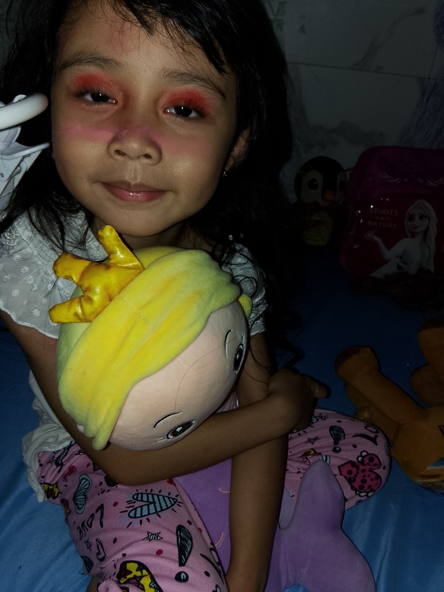 A young girl with dark hair and a slight smile is the central subject of this indoor portrait. She wears a white patterned top and pink pajama bottoms adorned with various playful graphics, including hearts, stars, butterflies, and the word "Love." Her face is notably covered with bright red-pink eyeshadow applied heavily to her eyelids, under-eyes, and extending across the bridge of her nose and cheeks, creating a dramatic, playful makeup look.
The girl cradles a plush doll with light skin, yellow hair or a hat, and a small golden crown. Her arms are wrapped around the doll, holding it close. In the background to her right, a dark blue surface, likely a bedsheet or blanket, serves as the setting. Several other toys are visible: a dark penguin stuffed animal, a brown plush toy, and a pink bag or backpack. The bag features an image of Elsa from Disney's Frozen, along with the partially visible text "SPIRITS of NATURE." A purple plush item, possibly a mermaid doll or tail, lies below the girl.
The lighting in the scene is dim and ambient, suggesting an evening or nighttime setting indoors. The overall mood is one of childlike playfulness and comfort, likely captured within a home in Davao City, Philippines.