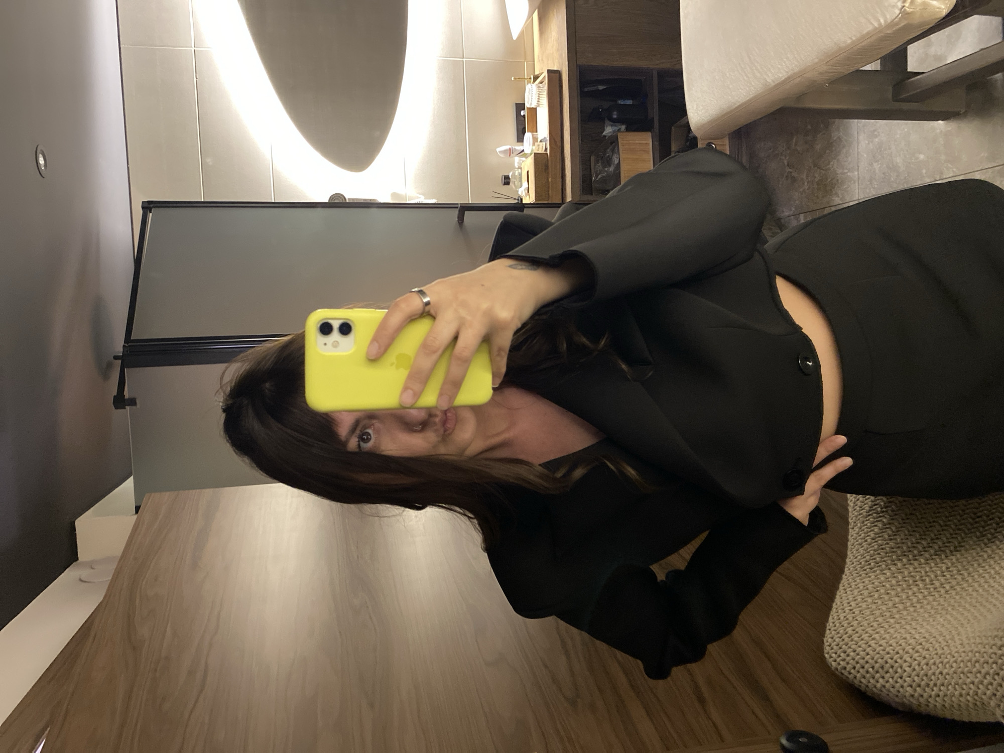 The image displays a mirror selfie of a woman within a modern interior, likely a bathroom or dressing room. The woman has dark hair with bangs and holds a bright yellow smartphone (iPhone) in her left hand, partially obscuring her face. Her visible right eye is dark, a nose stud is present in her right nostril, and her lips are slightly pursed. She wears a black, long-sleeved, V-neck cropped top, exposing a portion of her midriff, and black bottoms. A silver ring adorns her left ring finger, and a small dark tattoo is visible on her inner left wrist. Her right hand is placed on her hip. The background to her left consists of a light-colored wooden wall with visible grain, above a beige knitted textile. To her right is a bathroom setup, featuring a wooden counter holding a tray with various items including bottles and cotton swabs, a partially visible white sink, and dark wooden shelves beneath. The wall behind the counter is tiled with light-colored rectangular tiles. A large, oval-shaped mirror with integrated illumination reflects a bright, curved light. A dark-framed partition is visible in the upper right. The ceiling is dark with recessed lighting, and the floor is composed of dark-toned tiles. A padded bench with dark wooden legs is positioned on the far right.
