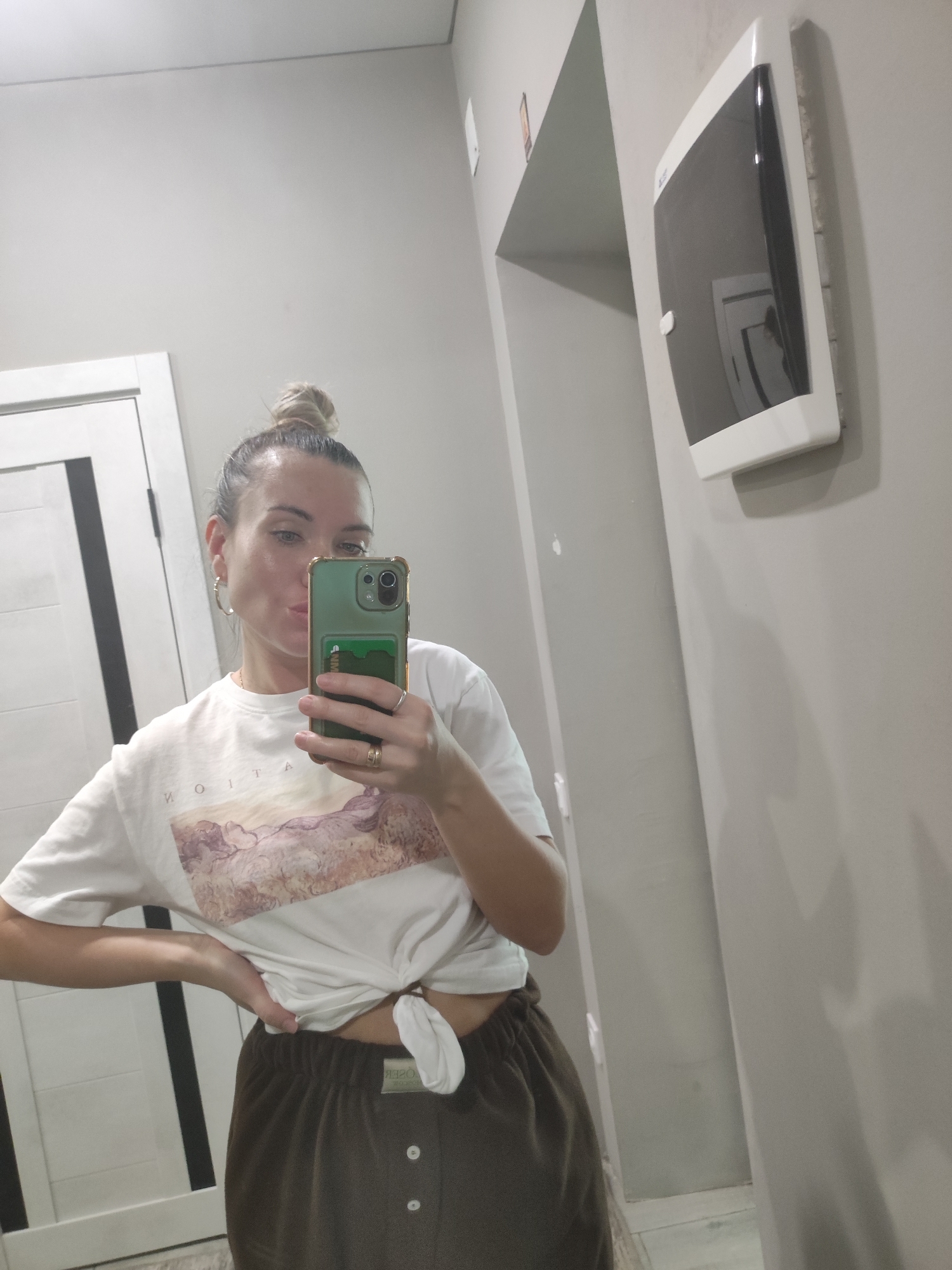 A woman with blonde hair in a high bun is taking a selfie in a bathroom. She is wearing a white t-shirt with a nature print and brown pajama bottoms tied at the waist. She is holding a green smartphone up to her face. The bathroom has a white door with black vertical stripes, light gray walls, and a white dispenser mounted on the wall. The location appears to be in a private residence in Votkinsk, Russia.