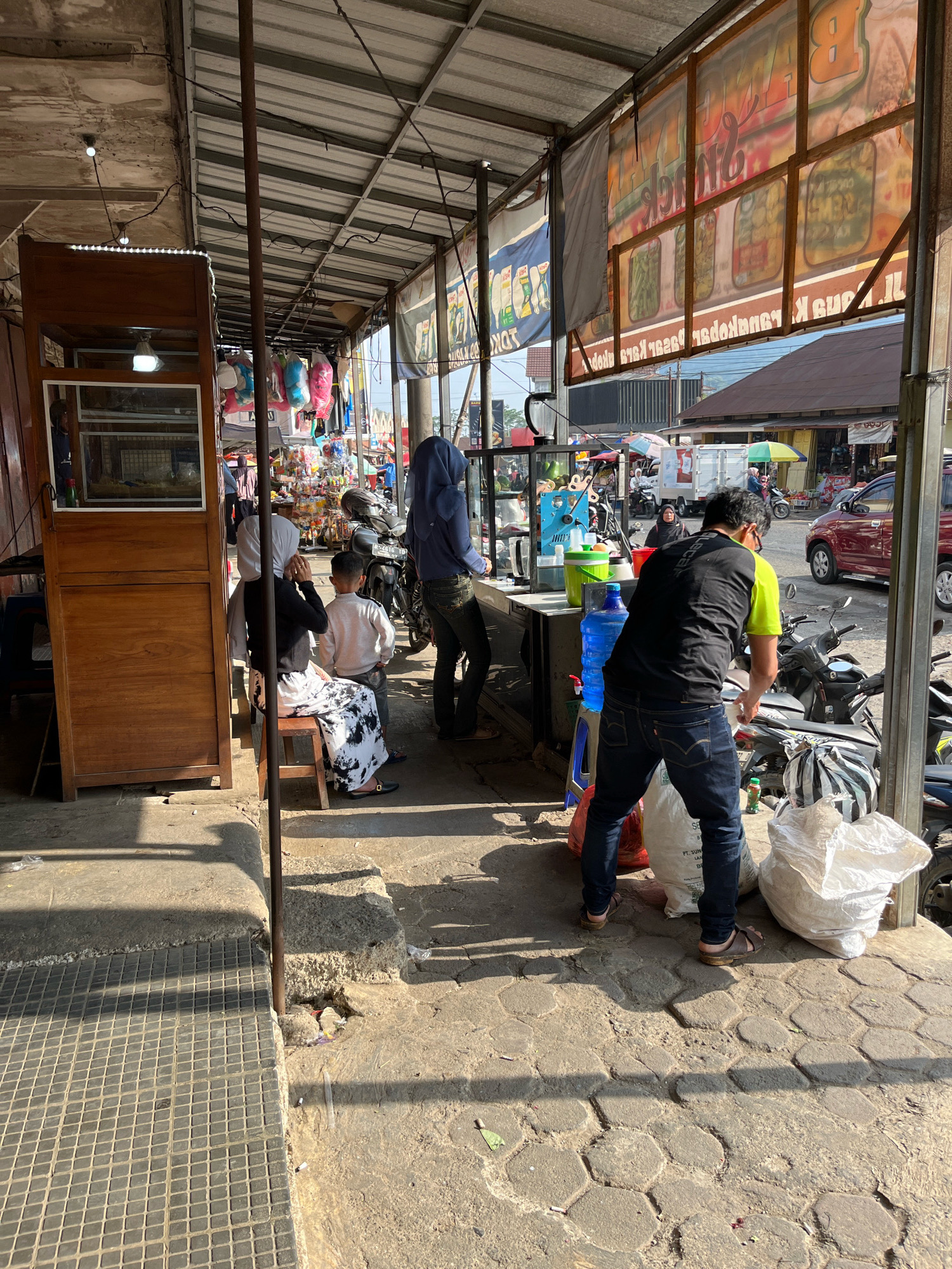 The image captures a street scene in Karangkobar, Indonesia, likely a market area under a covered walkway. In the foreground, a man wearing a black and neon yellow shirt and dark jeans is bent over, interacting with large white and red bags on the ground. He is wearing sandals. To his left, a blue water jug and a green container are visible on a stand. Behind him, several motorcycles are parked.
In the mid-ground, several people are visible. A woman in a dark headscarf and a white and black patterned skirt is sitting on a wooden stool next to a wooden display cabinet. A young boy in a light-colored sweater stands near her. Further back, a person wearing a blue hooded garment stands near a motorcycle. Various stalls with goods are set up along the walkway, with colorful items and signage in the background.
The environment consists of a concrete walkway with hexagonal patterned paving stones in some areas. The walkway is covered by a corrugated metal roof supported by metal poles. Sunlight casts shadows across the scene, indicating a bright day. In the background, buildings and a red car are visible.