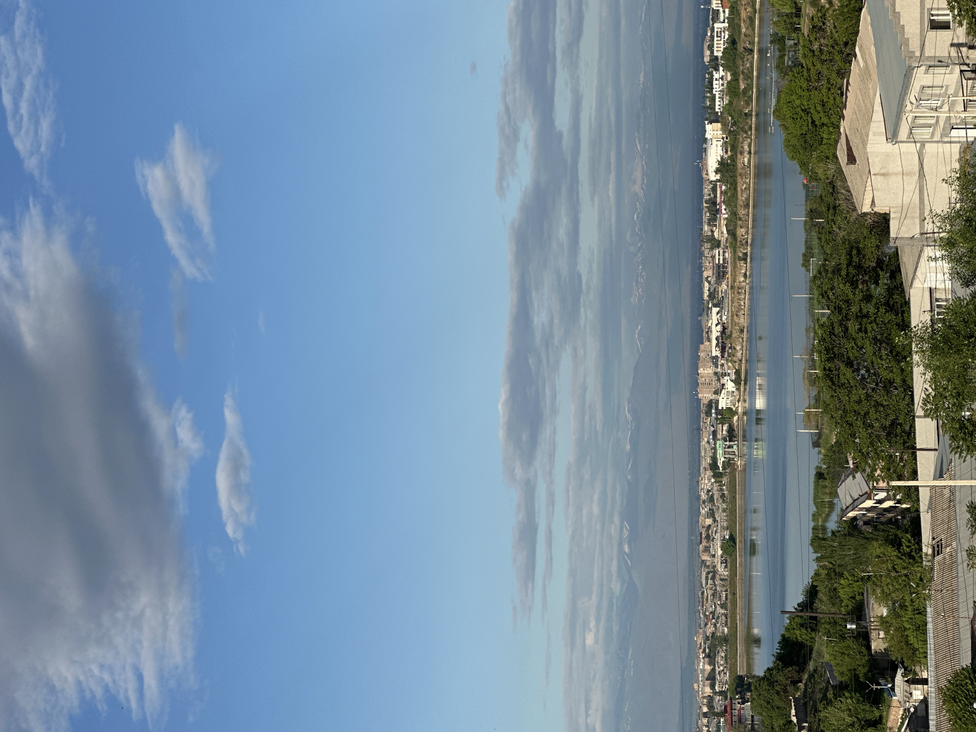 A vertical, wide shot shows Yerevan, Armenia with the blue sky and scattered clouds on the left. A calm water body reflects the city buildings, which are spread along the shore, separated from the right side of the frame by green trees and residential structures. The angle suggests a vantage point from within a building. The left side of the image displays an unblemished light blue sky, dotted with soft, white clouds. Towards the center, elongated, grey clouds hover closer to the skyline, mirroring the hazy outline of distant mountains that can be seen beyond the city. Power lines can be seen over the city.