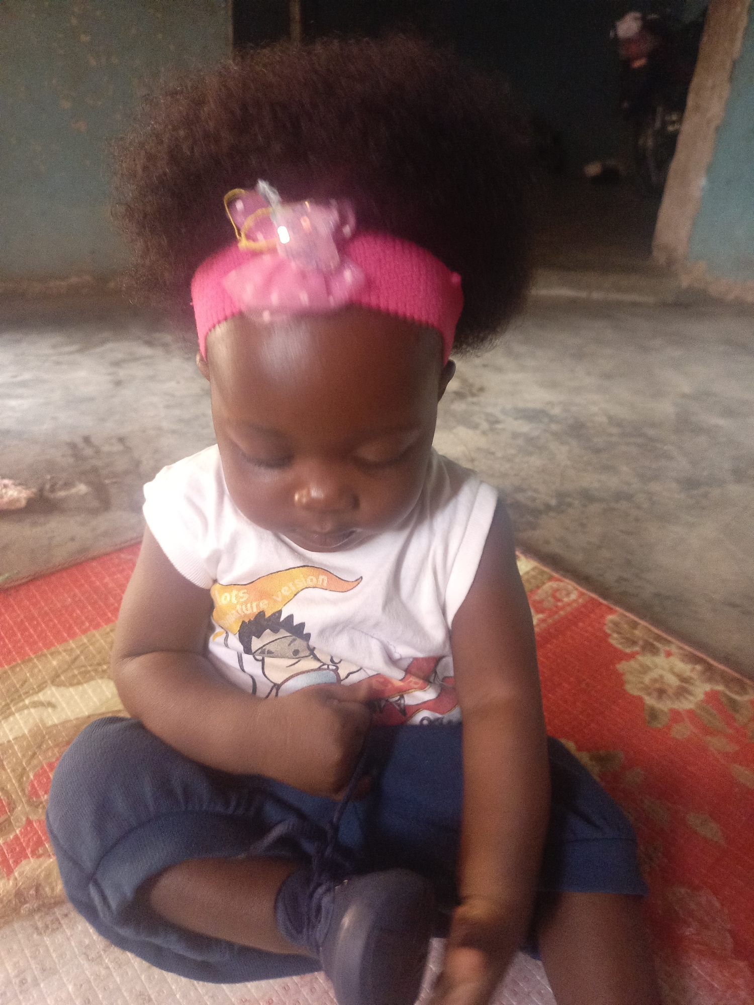 A young child with brown skin and dark curly hair, adorned with a pink headband featuring a bow, sits cross-legged on a patterned rug. The child wears a white T-shirt with a cartoon image and dark-colored pants, and appears to be looking down or focusing on something in their lap, possibly adjusting a dark shoe. The setting seems to be indoors, with a simple room visible in the background, including a partially visible wall and some objects further behind the child. The exact location is unknown.
