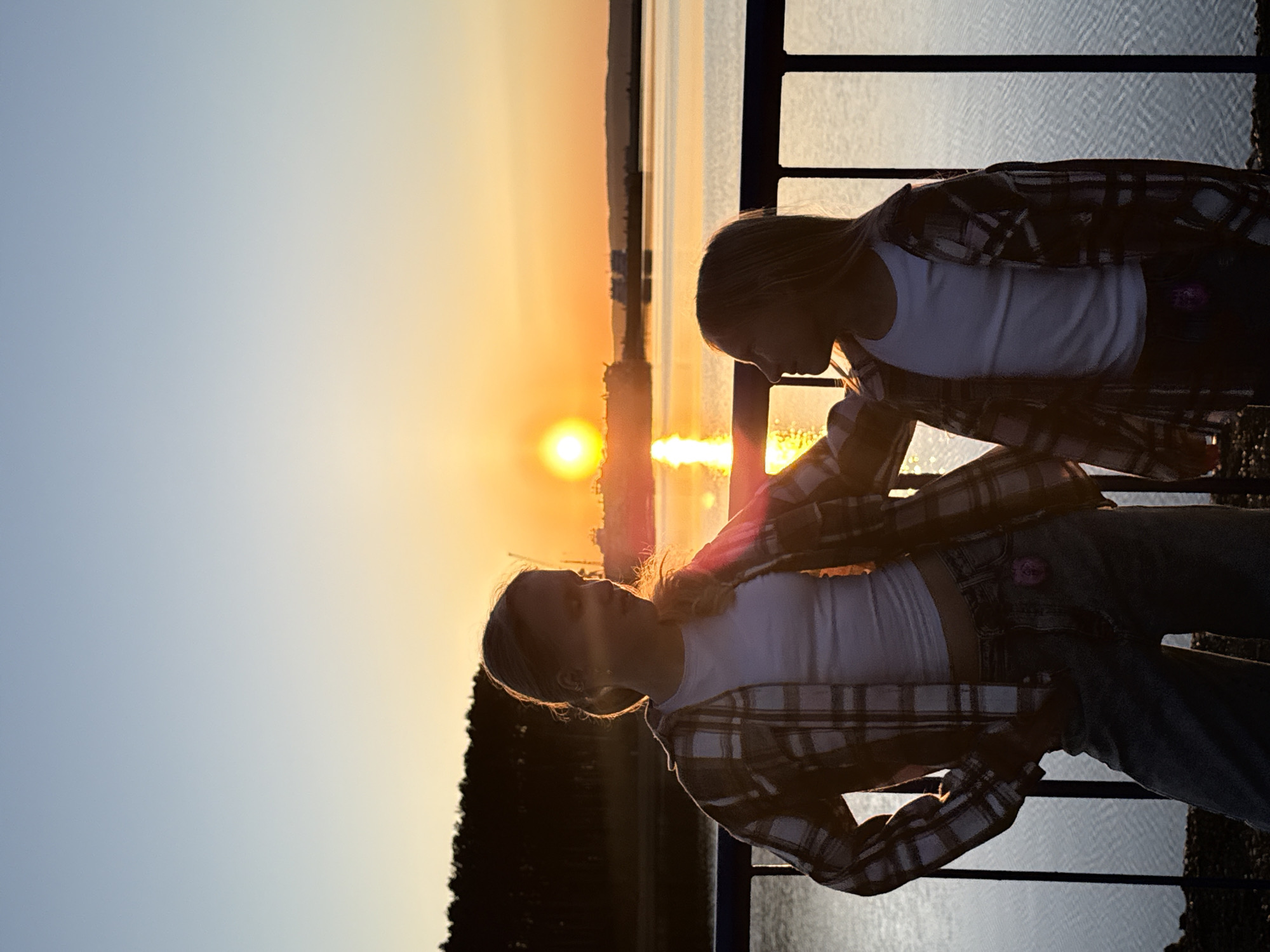 Two young girls are standing on a pier at sunset. They are wearing plaid shirts over white tops and jeans. The sun is setting over the water in the background, creating a golden glow. The location is likely near Chaikovsky, Russia, given the context provided. There is some sort of frame behind the girls, possibly a metal barrier, and trees in the background.