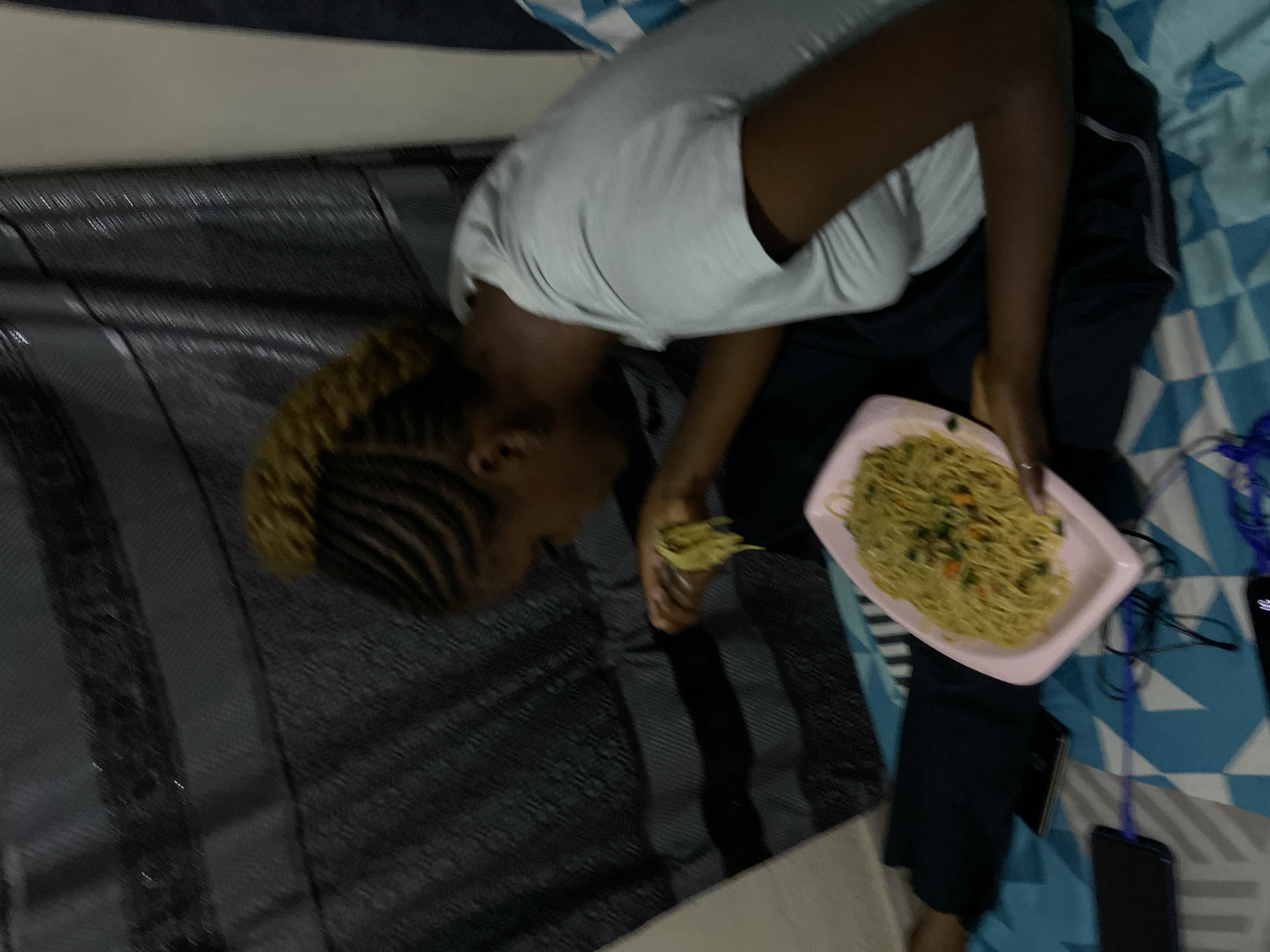 A young Black woman with braided hair in a high bun is sitting on a bed, eating a plate of noodles. She is wearing a white t-shirt and dark pants. The bed is covered with a blue and white patterned sheet. Behind her, there is a dark curtain with horizontal stripes. Two cell phones and some charging cables are visible on the bed. The scene appears to be in a domestic setting, likely a bedroom. The location is not specified in the image itself, and therefore cannot be definitively stated as Rafin Par, Nigeria.