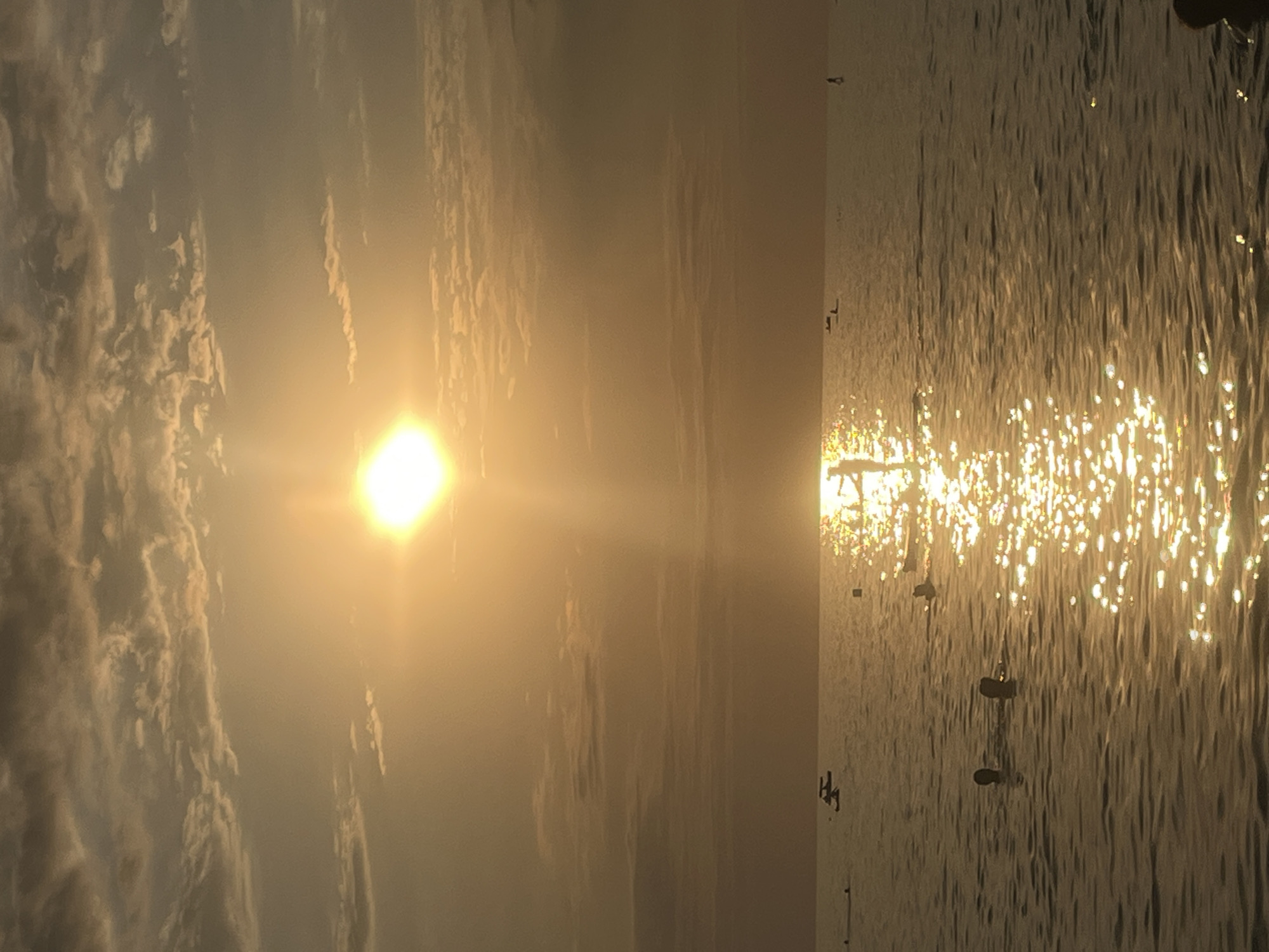 The image depicts a sunset over the sea, likely on the coast of Sochi, Russia. The sun, a bright, glowing orb, is positioned in the upper center of the frame, casting a warm, golden light across the sky and water. The sky is filled with scattered clouds, tinged with the orange and yellow hues of the sunset. The sea is calm, with gentle ripples reflecting the sunlight, creating a shimmering path. Several people are visible in the water, some swimming, others on paddleboards. In the distance, a lone figure stands on a paddleboard, silhouetted against the setting sun. The overall atmosphere is peaceful and serene, characteristic of a summer evening by the sea.