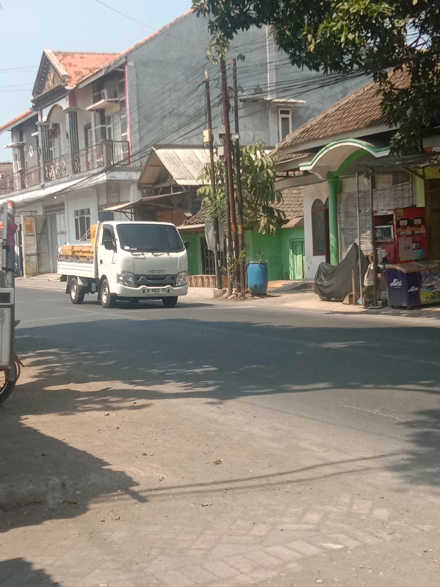 A white Isuzu truck is driving on a street in Karanganyar, Indonesia. The truck has a yellow bed and is carrying some cargo. The street is lined with buildings, some of which appear to be shops or residences. There are also trees and power lines visible. The sky is clear and the sun is shining, casting shadows on the street. The scene appears to be a typical day in a small Indonesian town.