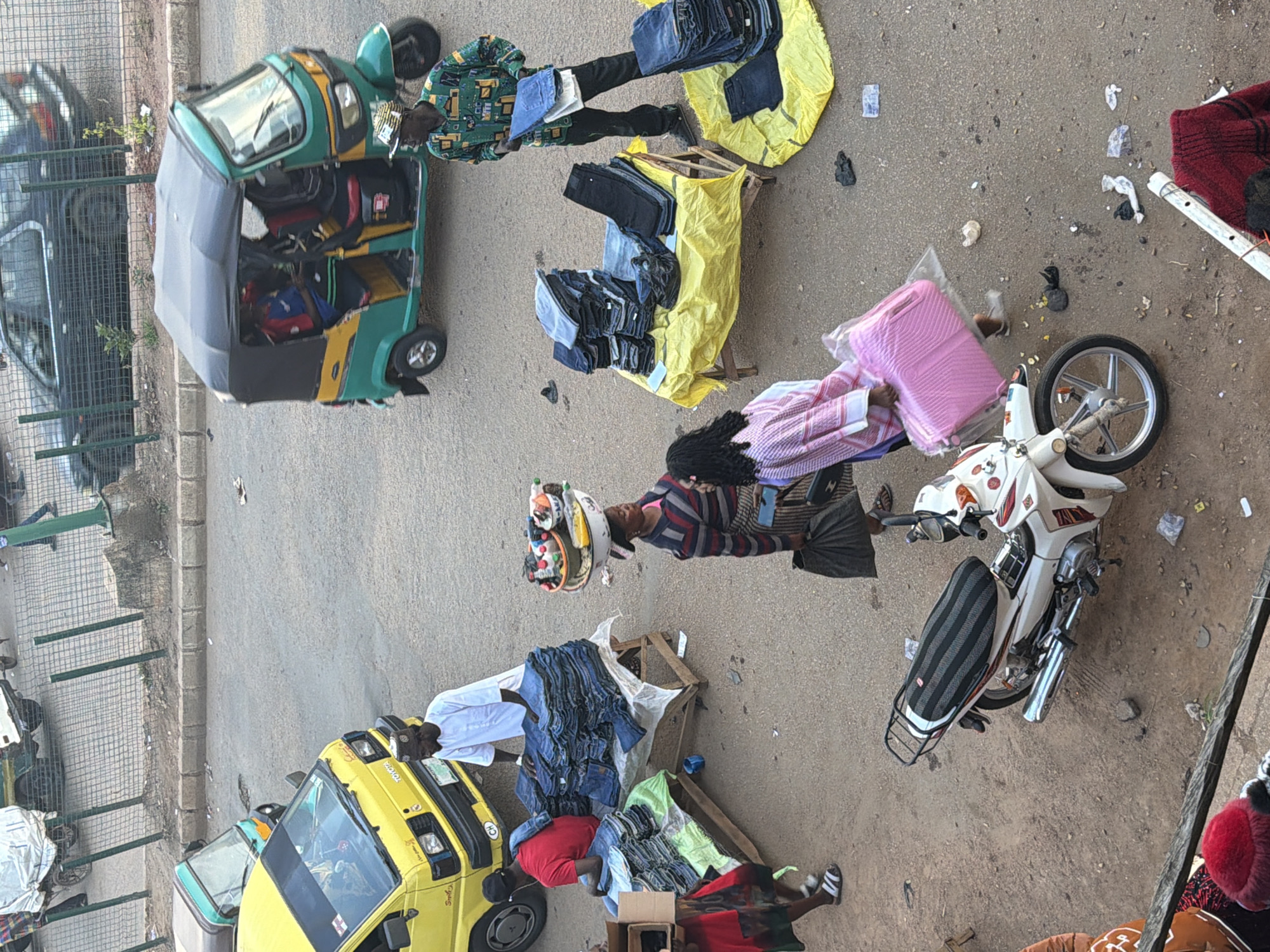 In Bukuru, Nigeria, an outdoor market scene unfolds with several individuals engaged in commerce and transit. A yellow Toyota van, with license plate CH, is parked on the asphalt road, alongside a green and yellow auto-rickshaw, also known as a "keke napep." Two men stand near a stall piled high with neatly folded blue jeans. One man, wearing a white thobe and skullcap, appears to be attending to the merchandise. The other, in a vibrantly patterned green shirt and black pants, holds up a pair of jeans. Nearby, a woman with a bowl of items balanced on her head, including plastic bottles and other goods, walks past. A second woman, in a pink and white striped outfit, carries a large pink suitcase. A white motorcycle is parked on the dirt and asphalt ground, its seat covered with a black and white striped fabric. The general environment suggests a bustling urban street market, with some debris scattered on the ground.