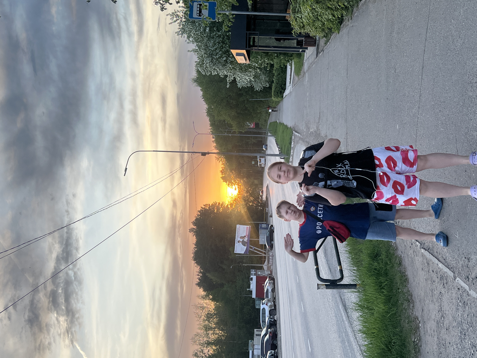 Two young boys are standing on a sidewalk next to a road. The boy on the left is wearing a blue shirt, denim shorts, and blue shoes. He has a red fanny pack slung across his chest. The boy on the right is wearing a black t-shirt, shorts with a red lips pattern, and purple shoes. The boys are both making hand gestures.
In the background, a sunset is visible between trees. Cars are parked along the side of the road. There is a bus stop sign on the right and a billboard on the left. The location is Chaikovsky, Russia.