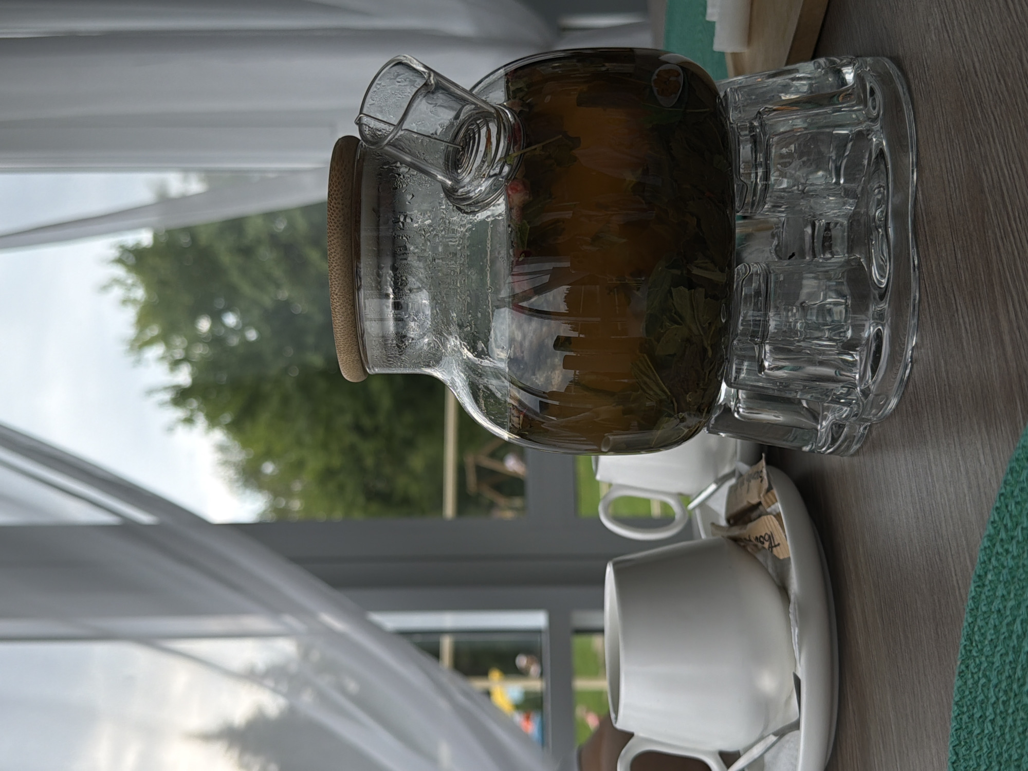 An indoor shot shows a glass teapot on a glass stand, filled with amber-colored liquid and tea leaves, positioned on a wooden table. To the left, a white teacup and saucer sit next to two sugar packets. In the background, sheer white curtains frame a window overlooking a garden with trees and a playground. The scene appears to be in the city of Chusovoy, Russia.