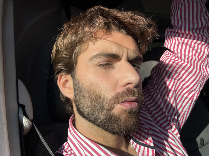 A male subject is depicted in a close-up portrait, located within the interior of a vehicle in Savigneux, France. The individual possesses brown, wavy hair and a full brown beard. He is wearing a long-sleeved shirt featuring vertical pink and white stripes, with a metallic chain visible at his neck. Strong natural light, consistent with direct sunlight, illuminates the right side of his face (viewer's left), causing a slight squint in his right eye. The left side of his face (viewer's right) and the vehicle's interior remain in shadow. A car seatbelt is visible across his left shoulder (viewer's right). The subject's right arm (viewer's left) is raised, with the striped sleeve prominently displayed. His head is slightly turned, and his lips are slightly parted.