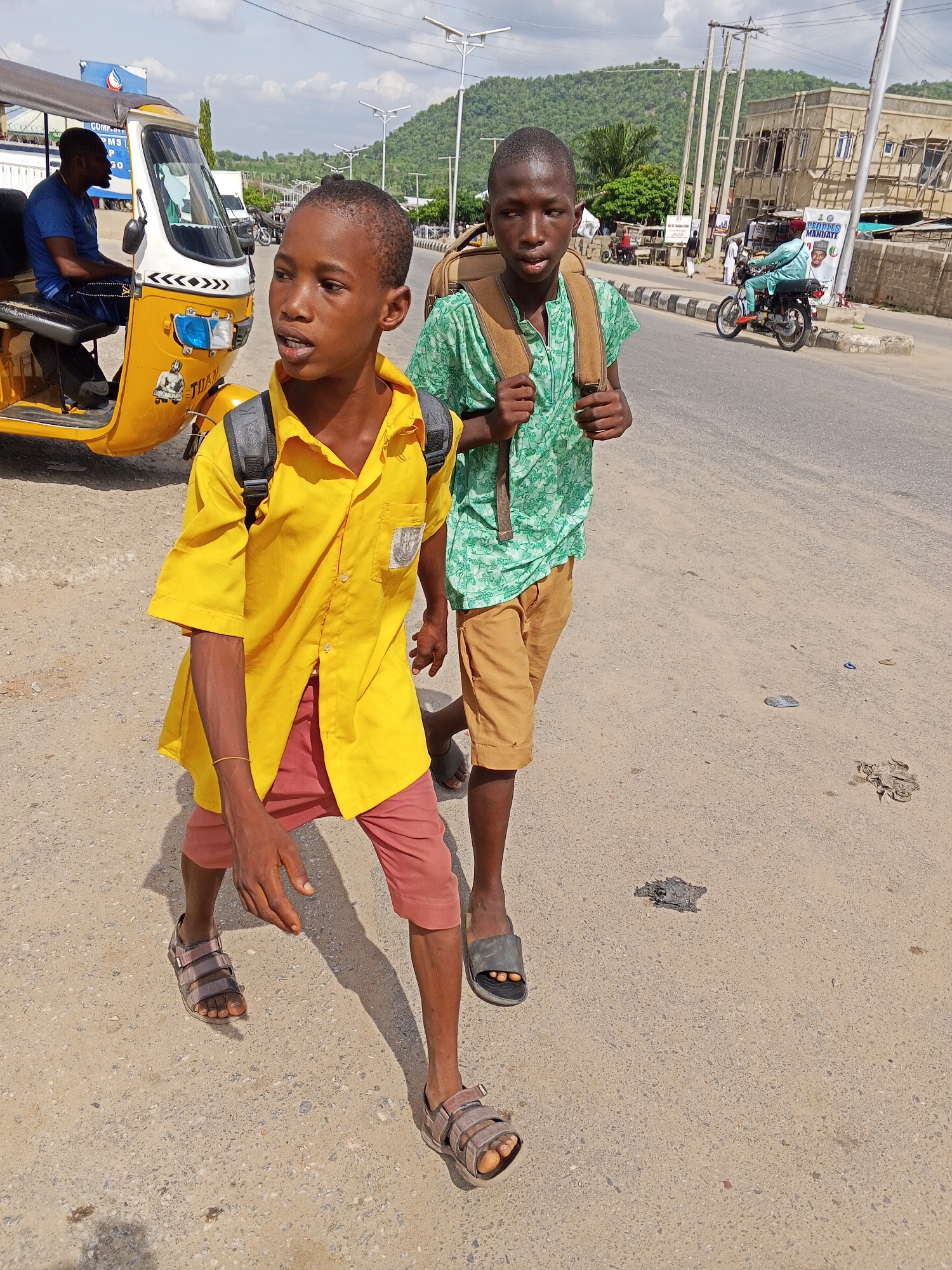 Two young boys in Zango-ndaji, Nigeria, walk on a paved road. The boy in the foreground wears a yellow, short-sleeved button-down shirt, red shorts, and brown sandals. He has a black backpack. The other boy wears a green, patterned, short-sleeved shirt, khaki shorts, black sandals, and a brown backpack. A yellow auto-rickshaw with a driver is parked on the left. A motorcycle is on the road further away. Buildings, utility poles, and a mountain are visible in the background. The sky is cloudy. The road surface has some debris.