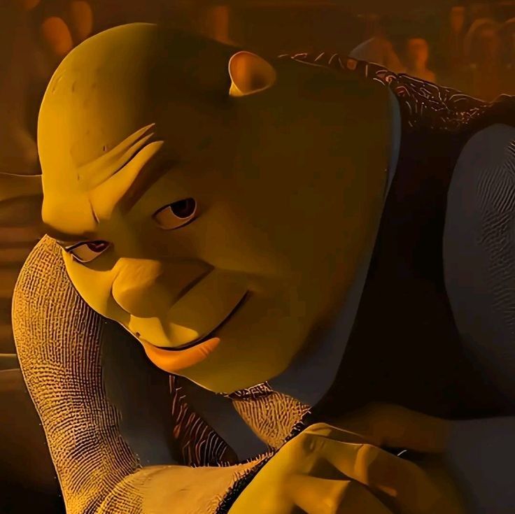 The image shows a close-up of a green ogre's face. The ogre is looking to the right of the image and smiling slightly. He is wearing a black shirt with a blue collar. The ogre appears to be in an indoor setting, with a blurred background. The ogre's facial expression is playful and slightly mischievous. The image is likely from a film or television show, and the ogre's character is likely fictional. The location in Geneng, Indonesia, is not relevant to the image as it is a fictional character.