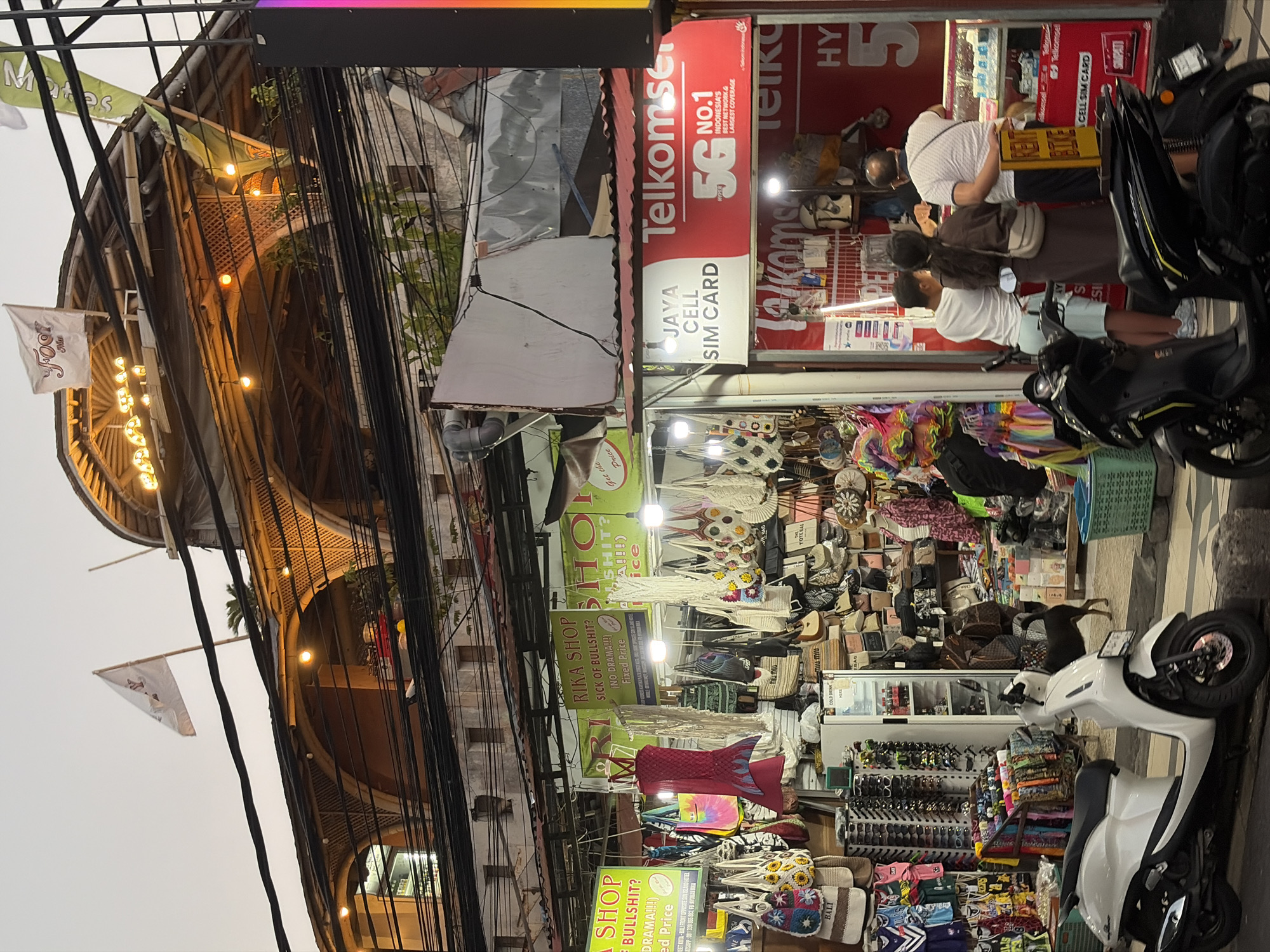 The image captures a bustling street scene in Kuta, Indonesia, likely during the late afternoon or early evening, judging by the ambient light and illuminated signs. The focus is on a row of shops selling various goods, including clothing, bags, and accessories. Overhead, a building with a distinct arched architectural style is visible, adorned with decorative lights and featuring a white flag with red lettering. A dense network of electrical wires runs across the upper portion of the frame, a common sight in many urban areas.
In the foreground, two scooters are parked on the pavement, one white and one black. A black dog is seen walking between the shops. Several people are engaged in shopping or interacting with store owners. One group of three individuals is standing near a "Telkomsel" cell phone shop, appearing to be customers. Another person is seen from behind, facing towards the shops. The shops themselves are densely packed with merchandise, with racks of clothing, hanging bags, and display cases filled with sunglasses. Signs in English and potentially Indonesian advertise "Rika Shop," "Sick of Bullshit," and "Jaya Cell SIM Card." A cooler labeled "Cold Drink" is visible inside one of the shops. The overall impression is one of a lively commercial area with a mix of local and tourist-oriented businesses.