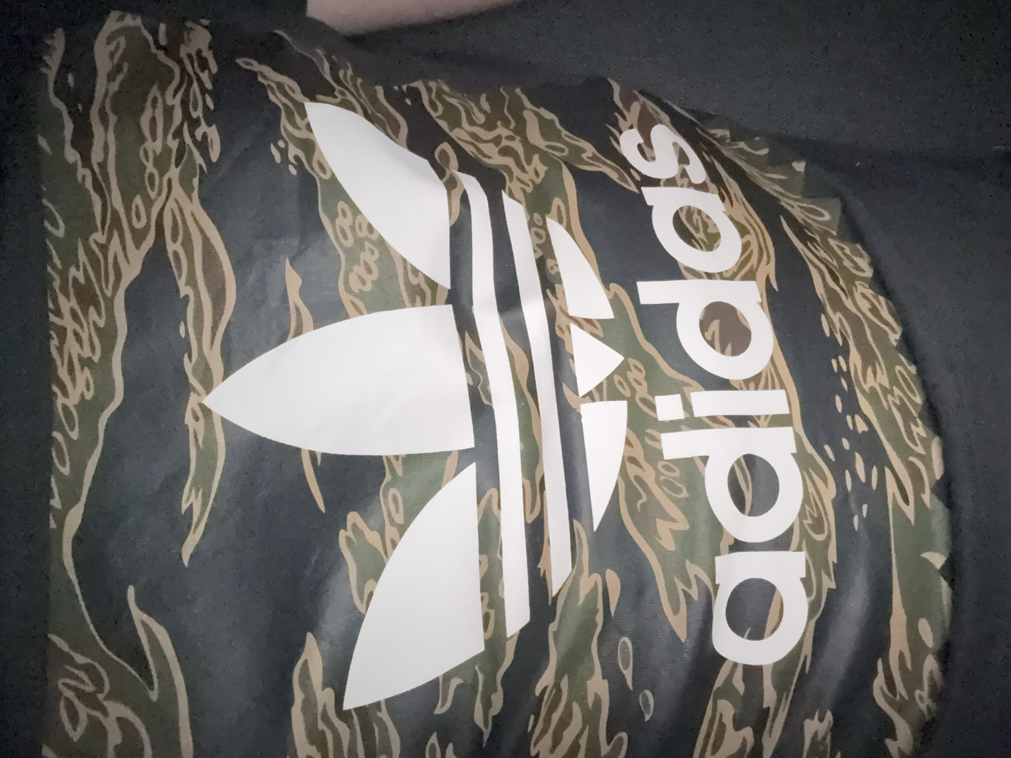 The image shows a close-up of an Adidas logo printed on a fabric with a camouflage pattern. The Adidas logo is white, while the camouflage pattern is a mix of green, brown, and black. The text "adidas" is printed next to the trefoil logo. The image appears to be taken indoors under dim lighting. The exact location in Stara Zagora, Bulgaria is unknown.