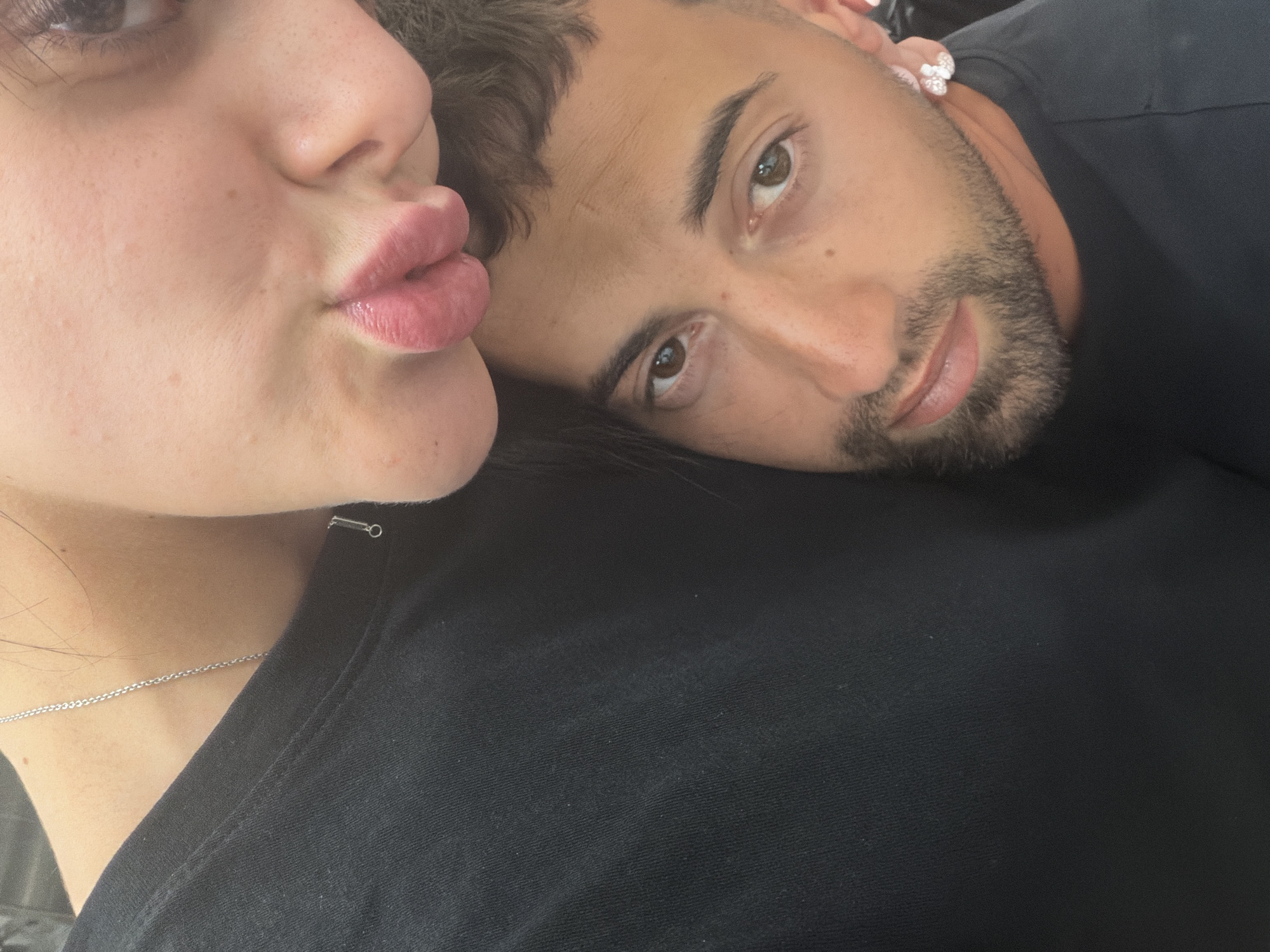 The image is a close-up, selfie-style shot of a couple. The woman's face is in the upper portion, with her lips puckered and close to the camera, obscuring part of her face. She is wearing a black t-shirt and a delicate silver necklace. The man's face is below hers, looking towards the camera. He has brown eyes, dark hair, and a beard. He is wearing a black t-shirt and a distinctive silver earring shaped like a flower. The background is mostly dark and indistinct, suggesting an indoor setting. There are no identifiable landmarks or environmental features that would indicate a specific location within the city of Plovdiv, Bulgaria. The image captures an intimate moment between the couple.