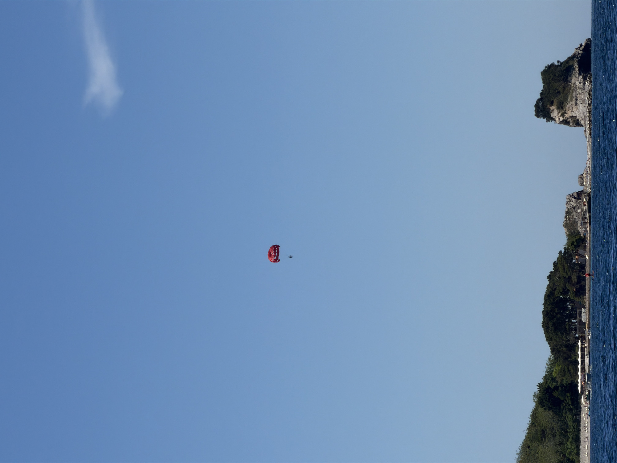 The image shows a bright, sunny day with a clear blue sky over the Adriatic Sea. In the distance, a red parachute, likely used for parasailing, is visible in the sky with two people suspended beneath it. The foreground features the coastline of Budva, Montenegro, with lush green trees and rocky formations. A small island with trees on top is situated to the right of the frame, and to the left, a strip of land with buildings and beach umbrellas can be seen. The water is a deep blue, and small boats are dotted around the coastline.