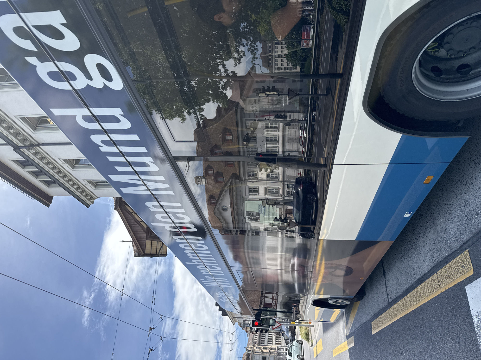 The image captures a side view of a modern, white and blue bus with large reflective windows, traveling on a city street. The bus has text in large white letters on its side, part of which is visible: "Wir nehmen den Mund ga". The reflection in the windows shows buildings with traditional architecture, typical of European cities, including a white building with ornate details and a building with a mansard roof. There is also a reflection of traffic lights and other vehicles. The street is paved, with yellow and white lane markings and a tactile paving strip. In the background, there are overhead wires for public transport and a bright blue sky with scattered clouds. A dark-colored car is visible in the reflection, driving on the road. The overall scene suggests a sunny day in an urban environment, likely in the city of Lucerne, Switzerland, given the architectural style and the presence of public transportation infrastructure.