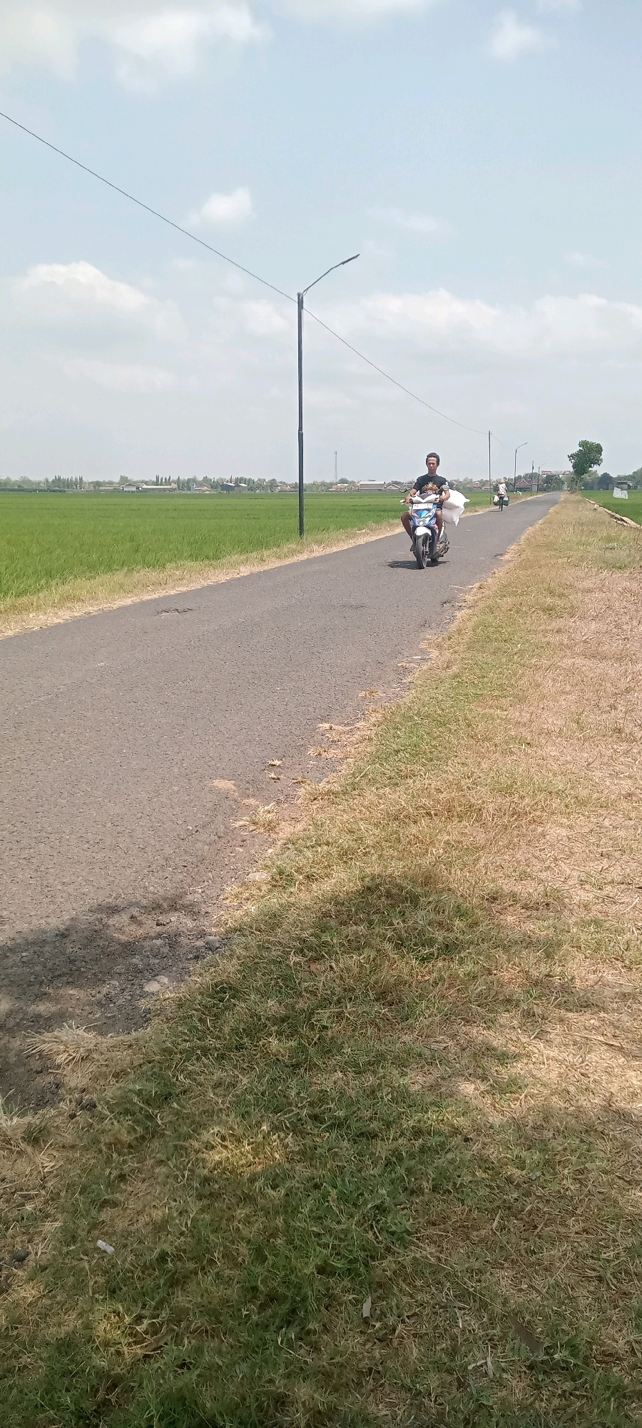 A man rides a motorcycle down a paved road in Ponowaren, Indonesia, with green rice fields on the left and dry grass on the right. He is wearing a dark t-shirt and is carrying two large white sacks on the back of the motorcycle. The sky is blue with scattered white clouds. A street light is visible on the left side of the road, and another street light is further down the road on the right. In the distance, there are houses and trees. The road appears to be in good condition, with some patches of wear. The overall scene suggests a rural or semi-rural setting on a sunny day.
