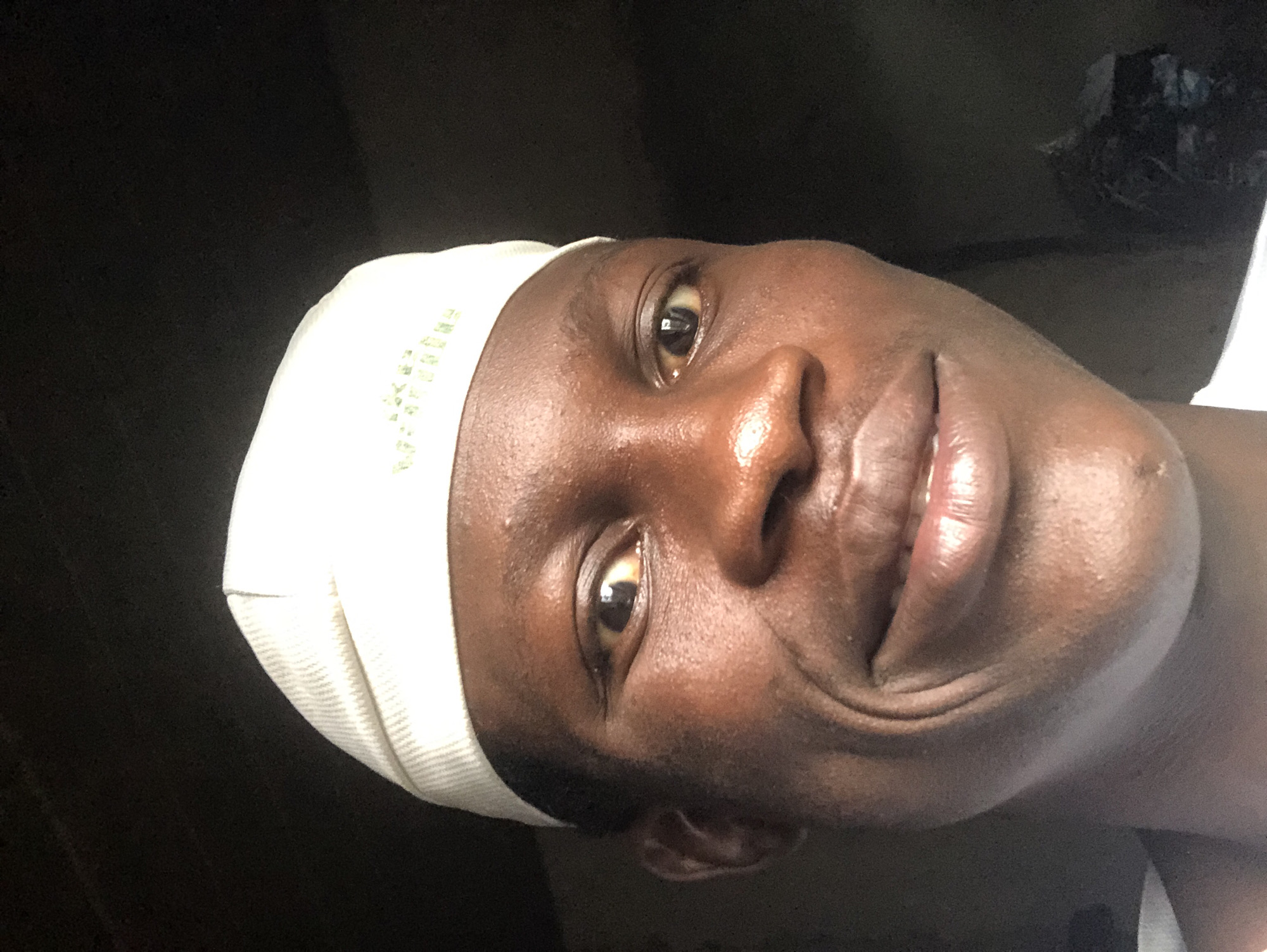 A close-up, head-and-shoulders shot, taken from a slightly low angle, shows a young Black male wearing a white, ribbed cap. The man's face is turned towards the camera and he is smiling, with his teeth slightly visible. His skin appears oily and reflects the light. The background is dark and indistinct, suggesting an indoor environment. Some faint green lettering is visible on the cap. The image is of moderate quality with some noise. The subject is likely in Ado, Nigeria, given the context provided.
