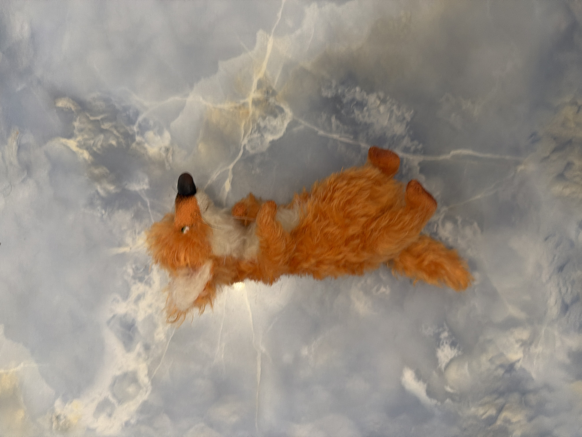 A stuffed fox toy is positioned vertically against a backdrop of blue and white marbled material. The fox, primarily orange with white accents on its ears and chest, is facing right. Its eyes are visible, and its snout is black. The toy's tail extends downwards. The marbled material features swirling patterns of lighter blue and white, creating a textured and abstract background.