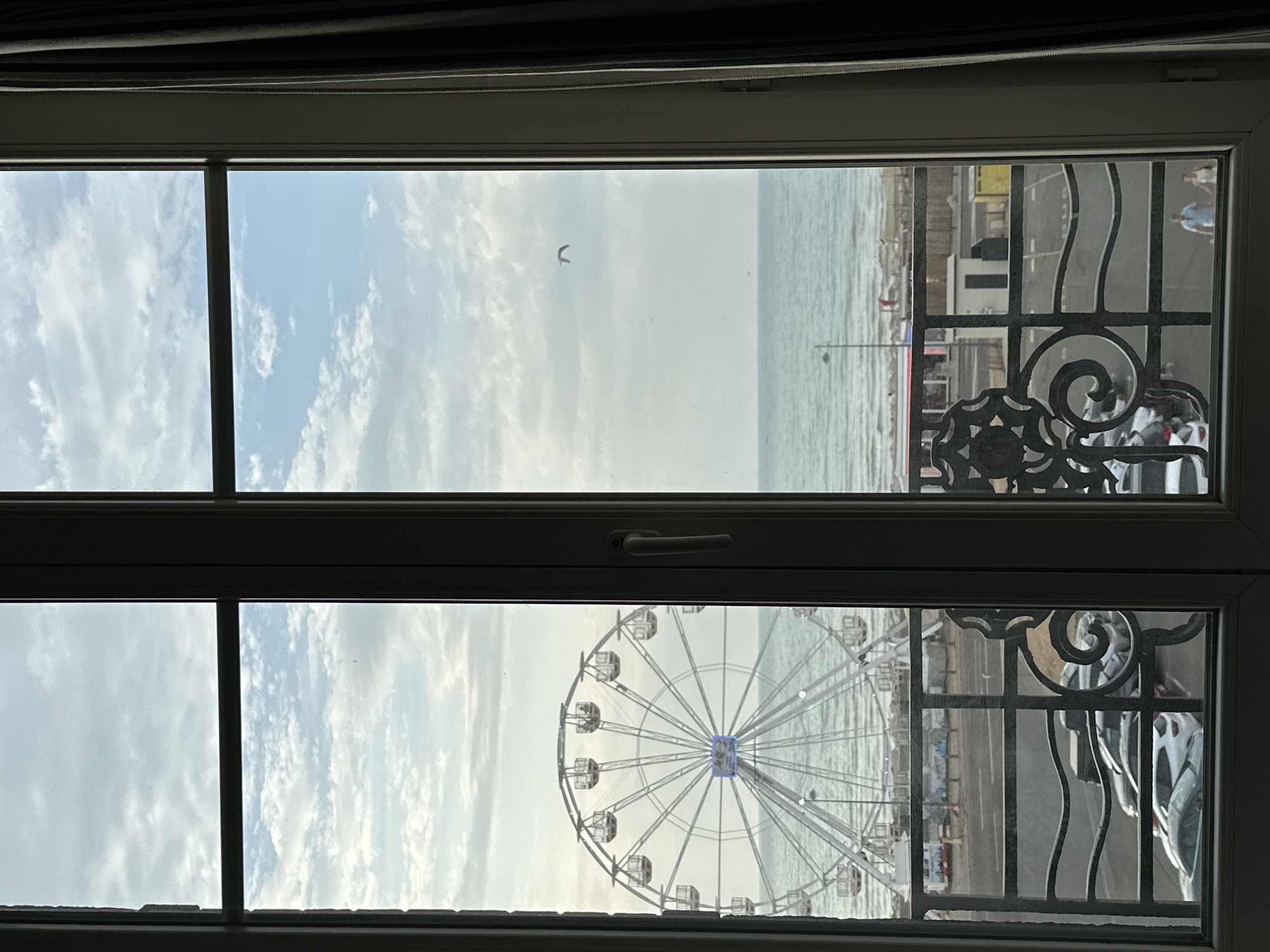 The image depicts a view through a window, overlooking the seafront of Le Tréport, France. In the foreground, a dark metal balcony railing with decorative elements is visible. Beyond the railing, a road runs parallel to the beach. Several parked cars are present on the road and in a parking area. Further in the distance, a large Ferris wheel dominates the left side of the frame, with its cabins and intricate structure clearly discernible. The sea stretches out beyond the Ferris wheel, with gentle waves rolling towards the shore. A beach area with a few scattered figures is visible along the coastline. The sky above is a mix of blue and white clouds. A single bird is captured in flight in the upper right portion of the sky. The overall scene conveys a coastal town atmosphere, likely during daytime with moderate cloud cover.
