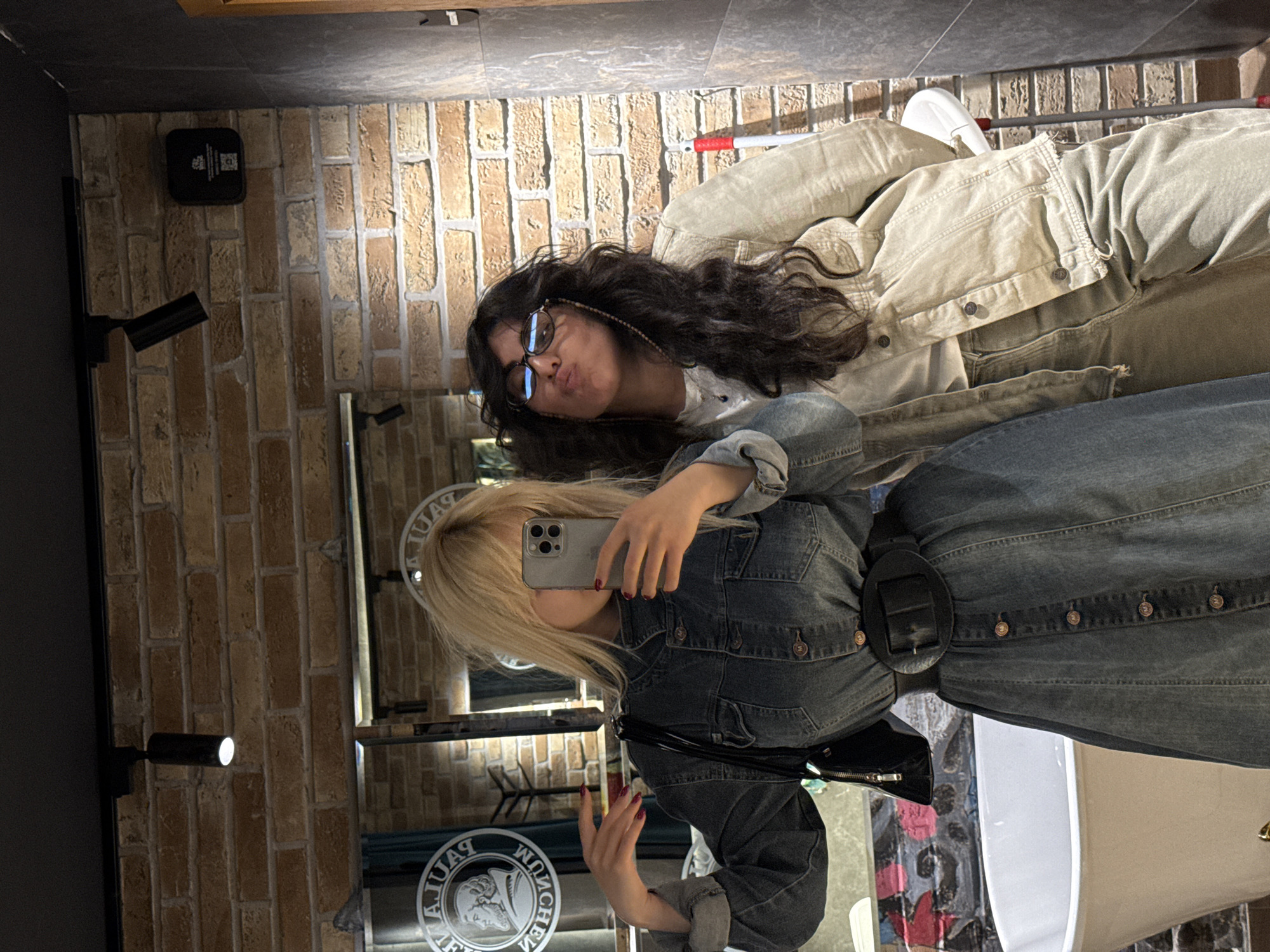 Two women are posing for a selfie in front of a mirror. The woman holding the phone has blonde hair and is wearing a denim dress with a wide belt and a black purse. The second woman has dark curly hair, is wearing glasses, and appears to be wearing a beige jacket. The background includes a brick wall, lighting fixtures, and the logo of "Parfumer." The location is likely a bathroom or dressing room in Yerevan, Armenia.