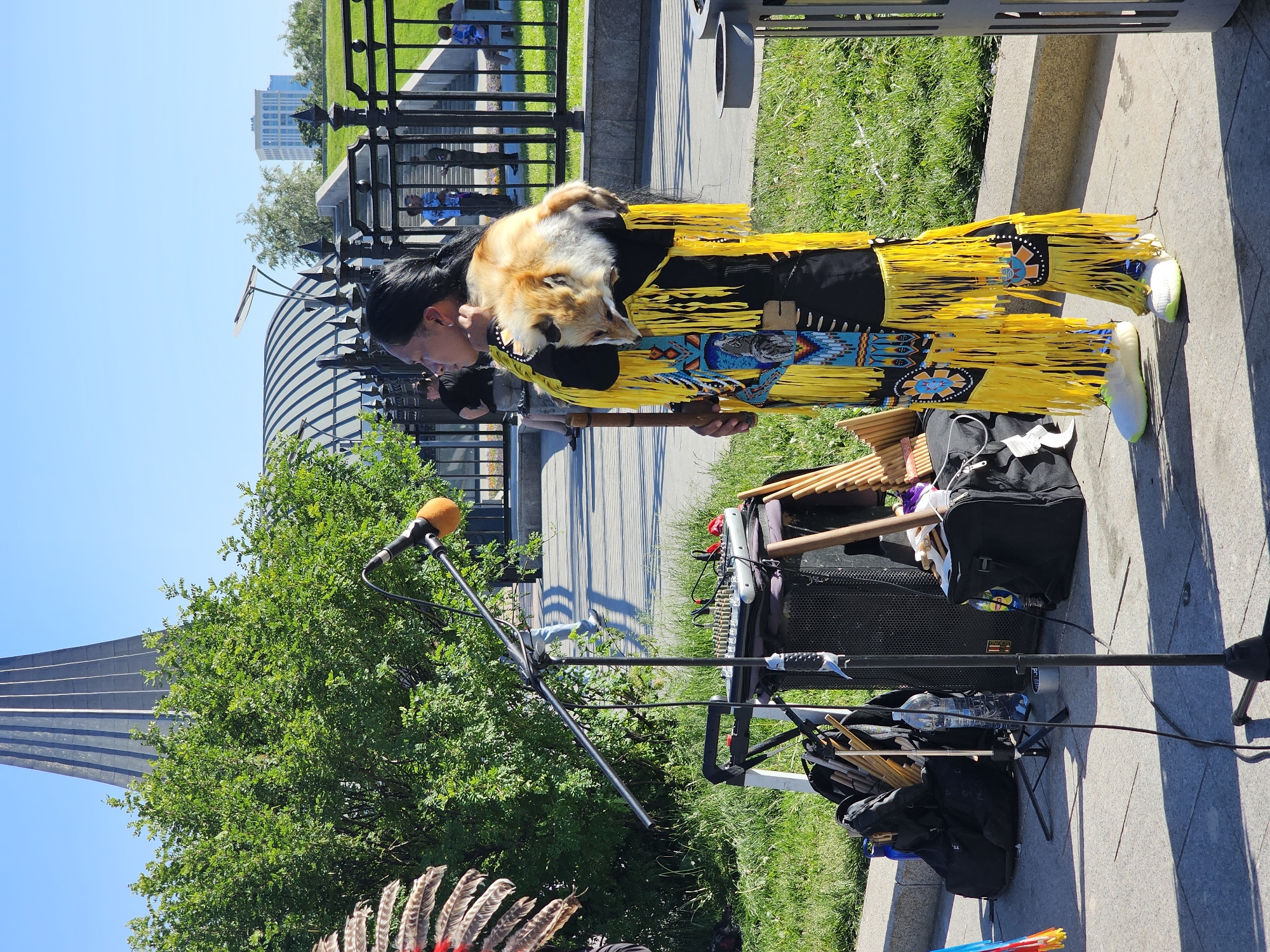 The image presents an outdoor scene under clear daylight, featuring a person in traditional attire, equipped for a performance. The central figure, with long dark hair tied back, stands facing left in profile. They wear a black garment richly decorated with bright yellow fringes and intricate beadwork featuring geometric patterns and a prominent eagle motif. Modern white and neon green sneakers are visible on their feet. A taxidermied fox pelt, including the head and fur, is draped over their right shoulder.
To the left of the individual, a performance setup is visible, consisting of a black microphone with an orange foam wind screen mounted on a stand, angled towards the performer. Beneath the microphone stand, a portable sound mixer and a black speaker rest on a support. Multiple pan flutes or similar tubular wind instruments are visible in bags and stacked around the equipment.
The background features a black ornamental metal fence. Beyond the fence, a paved area with steps leads up to a grassy slope, where several indistinct people are seen walking or standing. Lush green deciduous trees and bushes are abundant in the mid-ground. Against a clear blue sky in the upper left background, the distinctive dark grey, vertically ribbed conical structure of the **Monument to the Conquerors of Space** (also known as the Cosmonauts Alley Monument or Conquerors of Space Obelisk) is clearly visible. To the right of the monument, a large, arched, metal-domed structure and distant modern buildings further confirm the location as a public park setting in the city of Moscow, Russia.