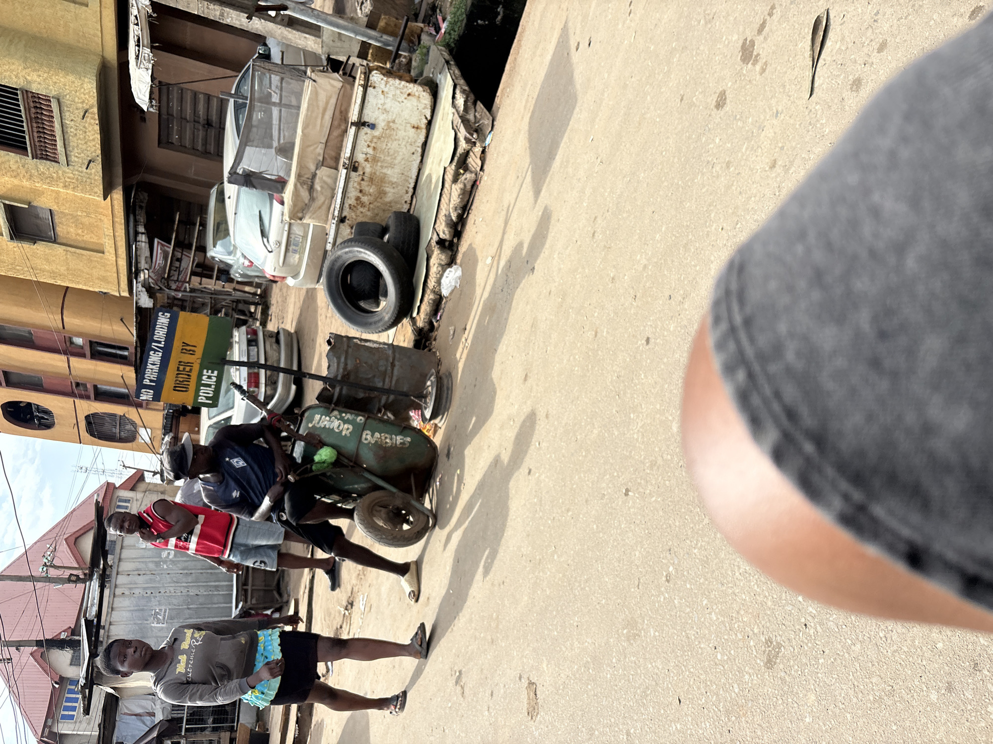 The image depicts a street scene in Onitsha, Nigeria. In the foreground, a person's leg and knee are visible, wearing dark-colored shorts or pants. The background shows a dusty, paved street with several people and objects.
On the left, a young woman with short hair, wearing a grey hoodie with text and a colorful ruffled skirt over black shorts, is walking towards the viewer. Behind her and slightly to the right, a man in a red sleeveless shirt and blue shorts stands with his hand on his chin.
In the center of the image, a man wearing a dark blue t-shirt and dark shorts sits on a green wheelbarrow. The wheelbarrow has text on its side that appears to read "JUNIOR BABIES". He is holding onto the handles of the wheelbarrow, which has one wheel visible. Next to the wheelbarrow, there are several tires, including two large ones stacked on top of each other.
Further in the background, a yellow building with balconies and a red-roofed structure are visible. A sign that reads "NO PARKING/LOADING ORDER BY POLICE" is mounted on a post. Several cars are parked or driving on the street, including a silver sedan and a white car. The environment appears to be a busy urban area with some debris on the ground. The lighting suggests it is daytime, with shadows cast on the street.