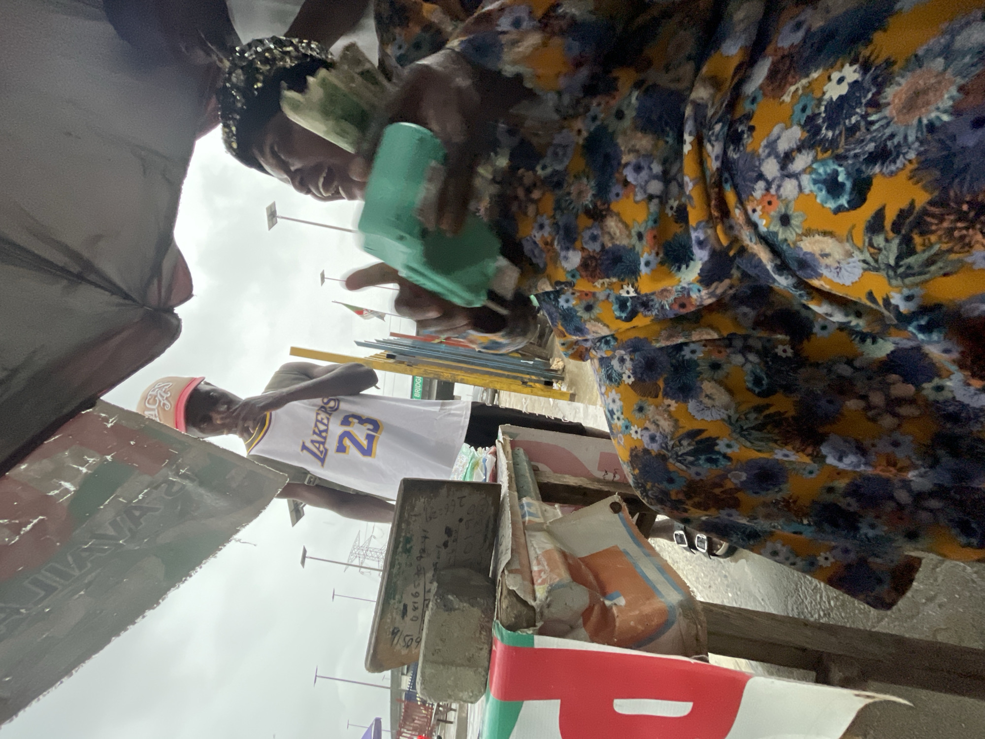 In Lagos, Nigeria, a young man wearing a white Los Angeles Lakers jersey with the number 23 and a red and beige baseball cap stands near a makeshift stall. He is looking towards a woman seated at the stall, who is wearing a vibrant floral-patterned dress and a sparkling headpiece. The woman is actively engaged with a small, bright teal-colored device, holding it in her right hand. She appears to be in the process of a transaction or interaction involving the device and some cash or other items in her hand. The stall itself is cluttered with various objects, including what appear to be bricks or construction materials, and possibly signage. The background suggests an outdoor, urban environment with overcast skies, and street infrastructure like lamp posts and a bridge visible in the distance.