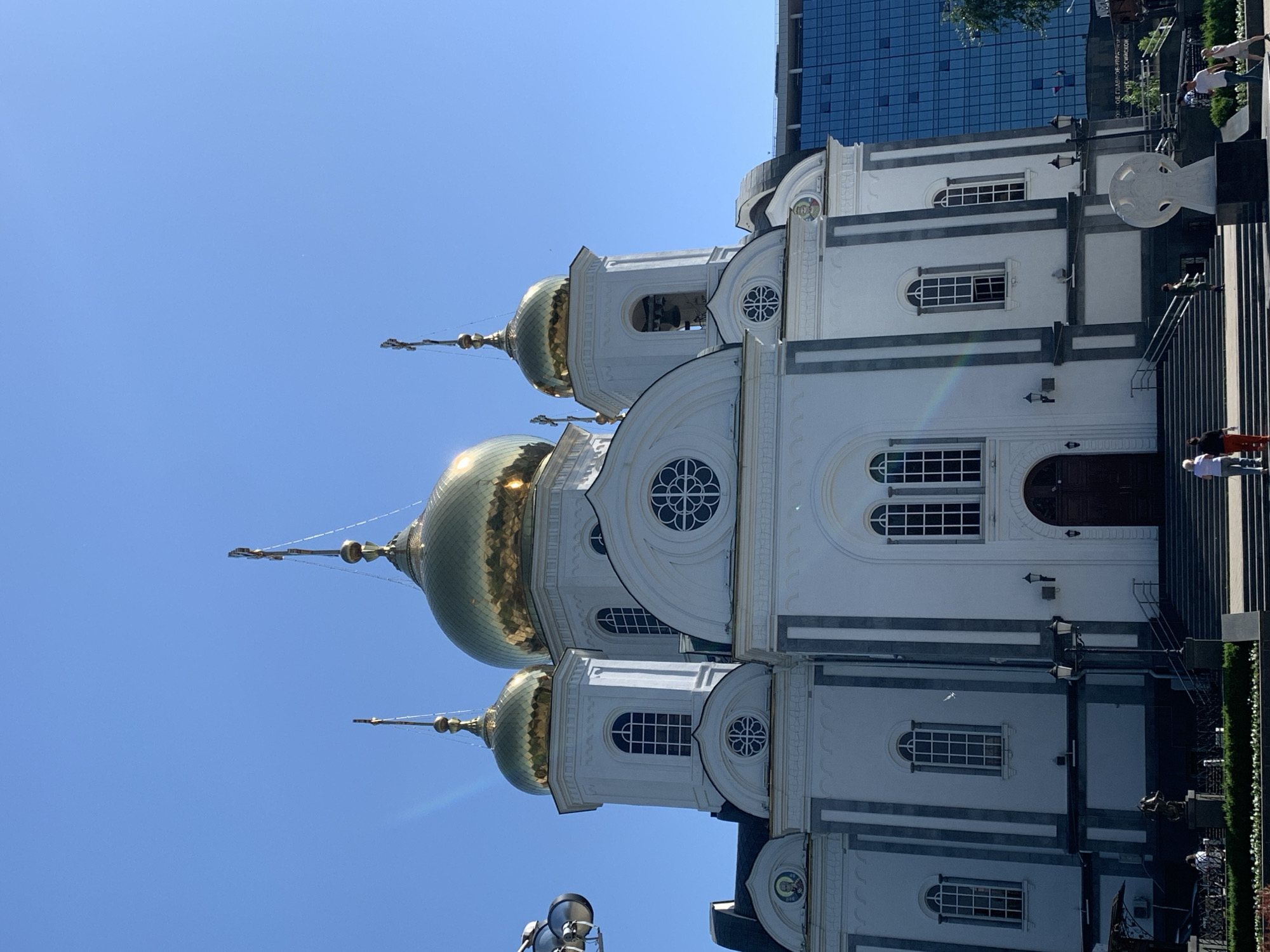 Here is a description of the image:
The image shows a white, ornate church with golden domes against a clear blue sky. The church has multiple levels, with arched windows and decorative elements. A modern building can be seen behind the church. People are visible near the church's entrance, and landscaping borders the building. The location appears to be an urban area.