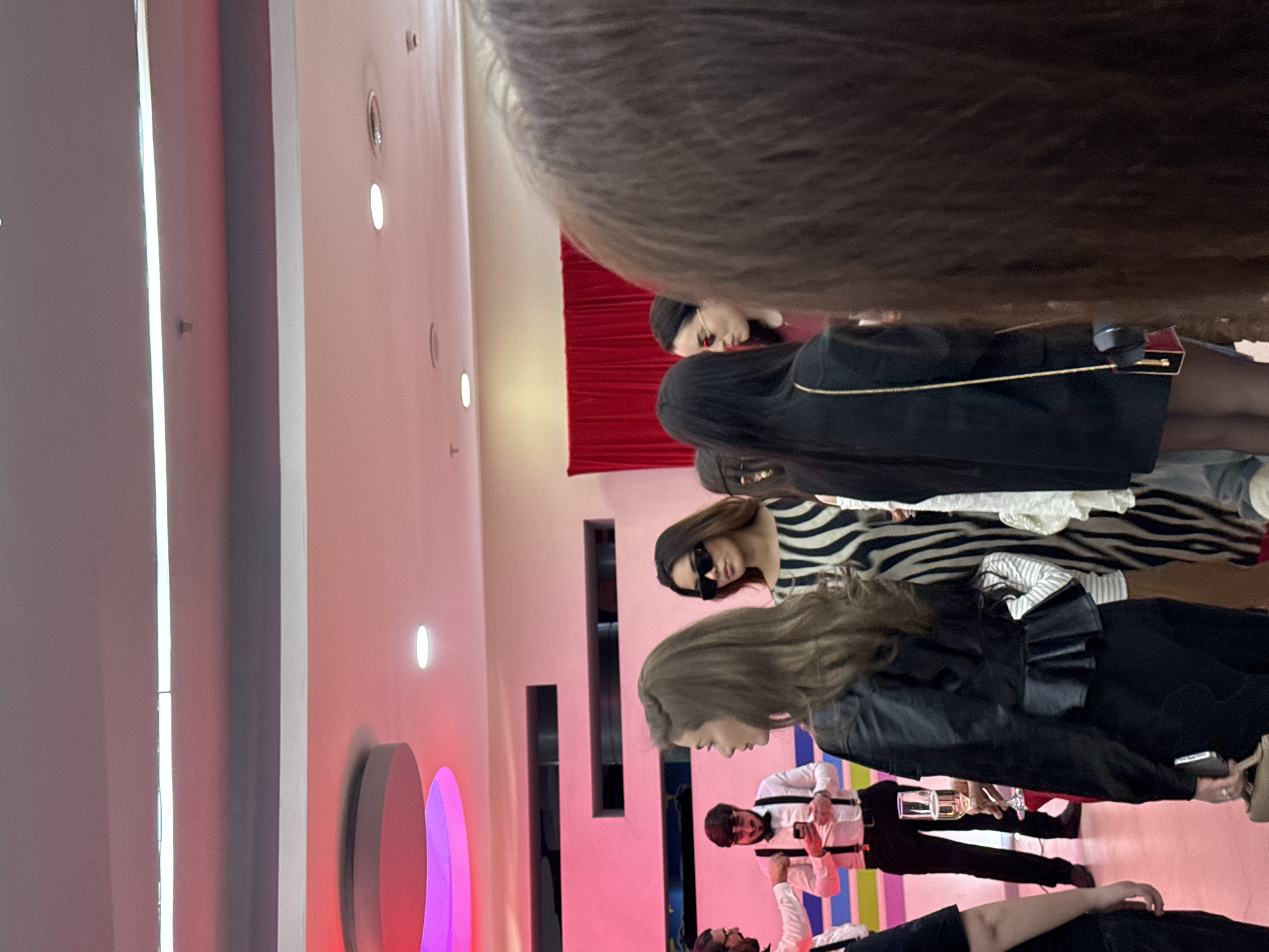 The image depicts a fashion show setting, possibly in Yerevan, Armenia, with several models wearing different outfits. One model is wearing a black and white zebra-striped dress, while another is in a black outfit with a leather jacket. Some of the outfits appear to be displayed on a runway against a backdrop of red curtains and pink walls with vertical decorative elements. There are photographers and other personnel present, some holding champagne glasses. The event appears to be well-lit. The foreground is partially obscured by the back of someone's head.