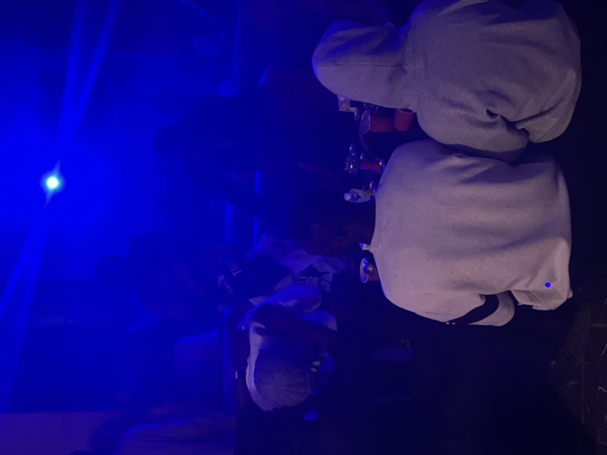 The image depicts a dimly lit indoor environment, saturated with blue lighting, suggesting a night-time or club-like setting. Two individuals are prominently featured in the foreground, viewed from behind. The person on the left wears a light-colored sweatshirt with dark stripes on the sleeves. The person on the right is wearing a grey hooded sweatshirt. In the background, other figures are silhouetted against the blue light, some appearing to be seated or gathered around a surface. There are red cups and what appears to be a hookah setup on a table in the mid-ground. The overall atmosphere is hazy and suggestive of a social gathering. The specific location within Durimi, Nigeria, cannot be determined from the visual information provided in the image.