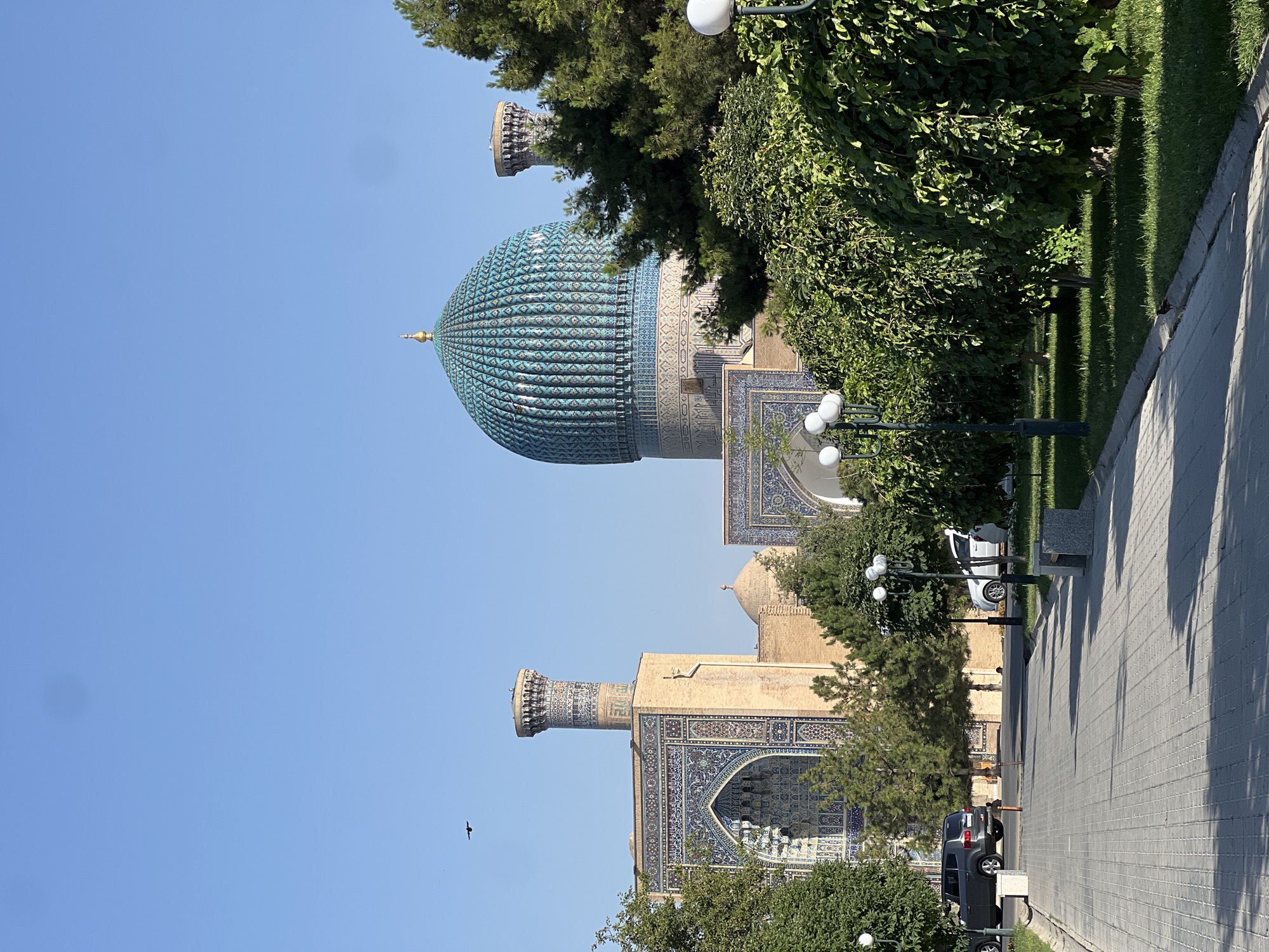 A wide-angle, eye-level shot captures a sunlit pathway in Samarkand City, Uzbekistan, leading towards a grand architectural complex featuring minarets and a prominent turquoise dome. The pathway, paved with grey interlocking bricks, is bordered by a meticulously manicured lawn on the right, with several rounded, leafy trees and modern streetlights. On the left, the pathway is also bordered by a lawn, with a grey SUV parked on the side. In the background, the architectural marvels of Samarkand are visible. The tallest structure is a minaret adorned with intricate blue and white geometric patterns. Adjacent to it is an arched entrance with similar detailed tilework. The most striking feature is the large, fluted turquoise dome of a mausoleum, topped with a golden finial, standing regally against a clear, pale blue sky. A smaller, lighter dome is visible behind the main structure. The scene suggests a peaceful, well-maintained public space with a focus on historical preservation and natural beauty. A bird is seen flying in the upper left quadrant of the sky.