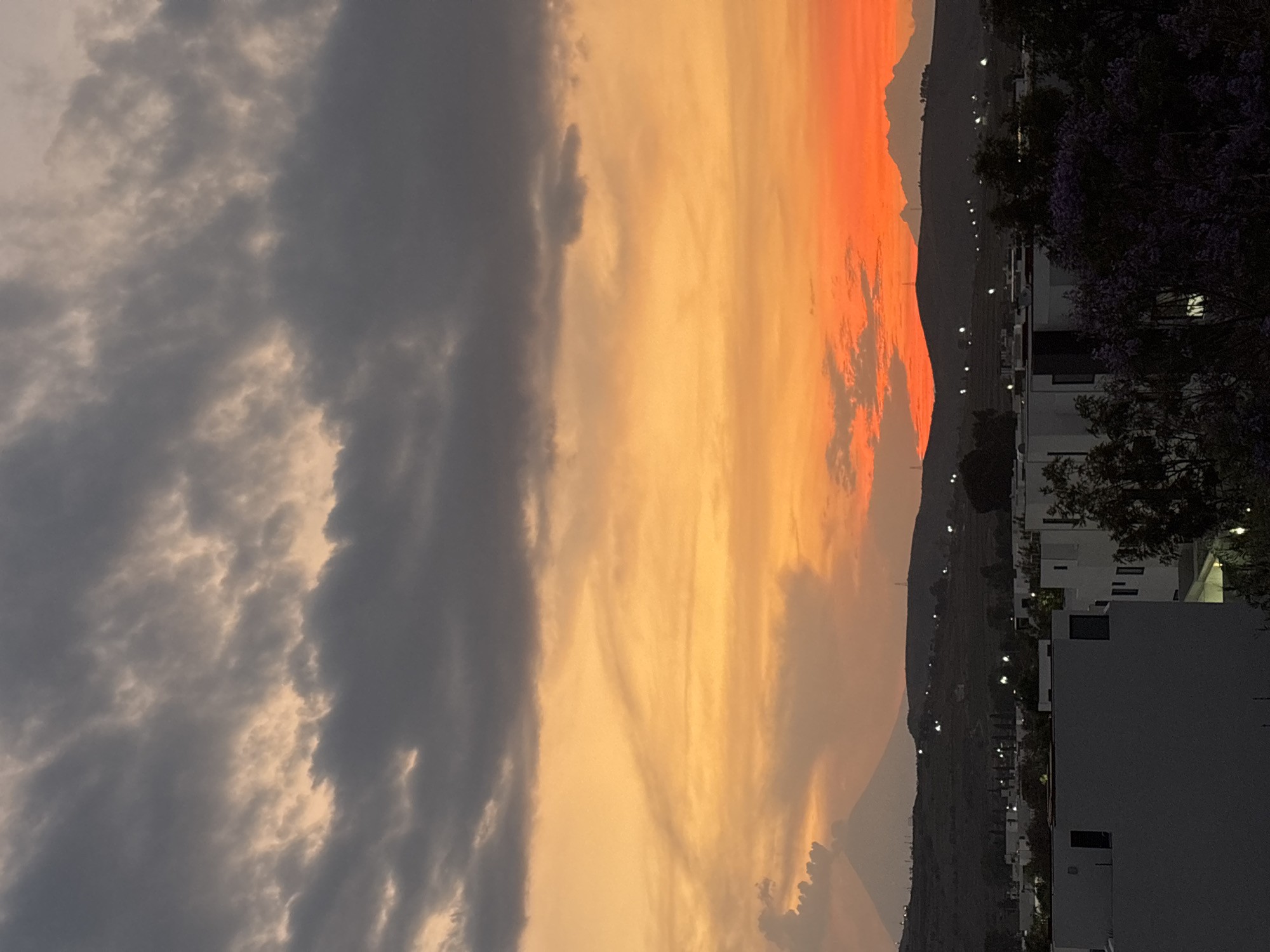 The image captures a sunset scene in Santa Clara Ocoyucan, Mexico. The sky is divided into three sections: the left side is covered with gray clouds, the center exhibits a blend of yellow and orange hues, and the rightmost part showcases a bright orange glow.
In the background, a mountain range is visible. Several houses and trees are also present, illuminated by streetlights. A building with a square architecture stands out on the right side.