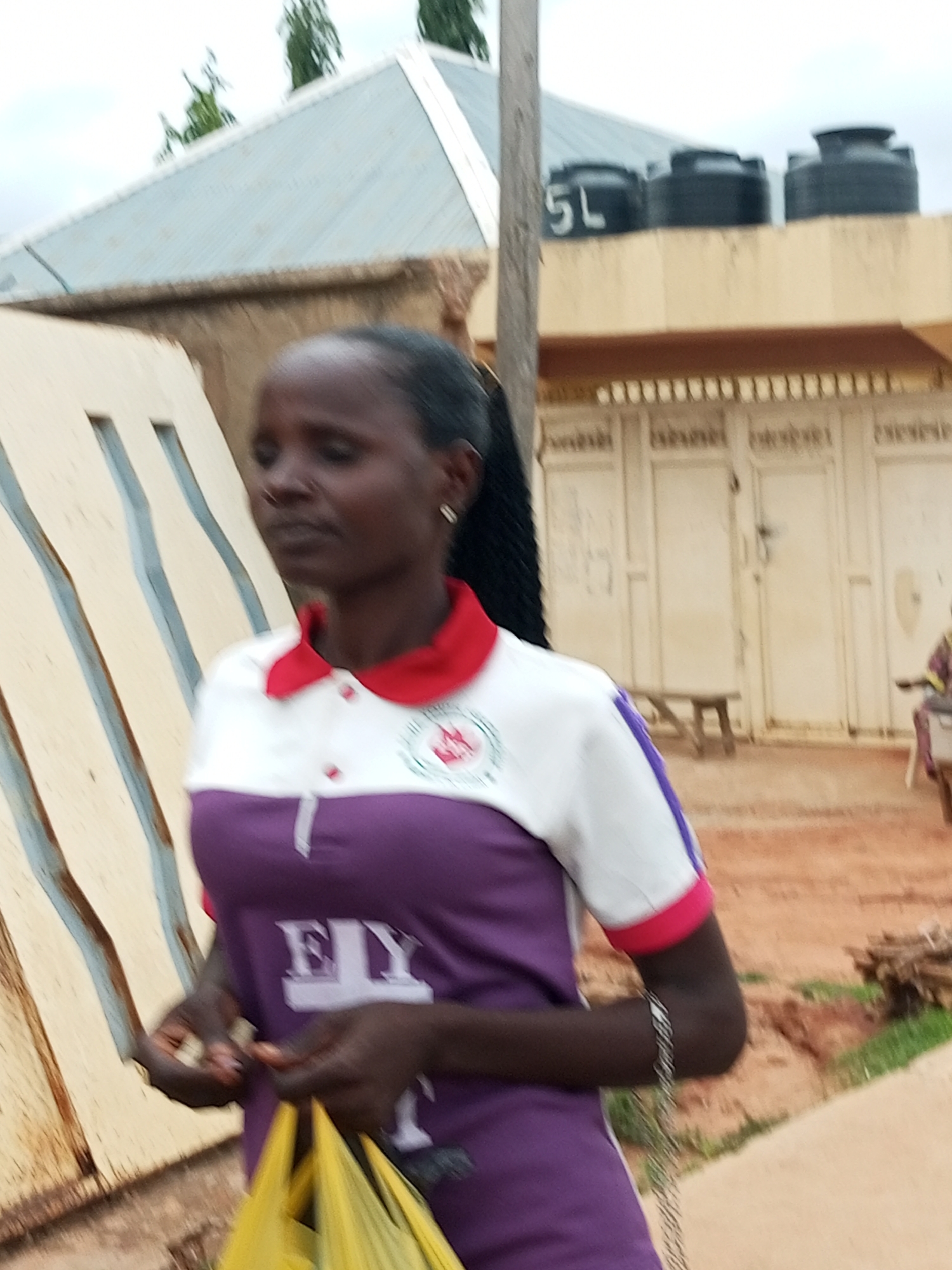 The image depicts a young woman standing in front of a building with a light-colored corrugated metal roof and beige walls. Three black water tanks are visible on top of the building. The woman is wearing a white shirt with purple accents and a red collar, and she is holding a yellow plastic bag. Her hair is styled in a long ponytail. The ground is reddish-brown, and there is some vegetation and wood debris in the background. A chain or bracelet is visible on her wrist. The scene appears to be in an outdoor environment in Tempure, Nigeria.
