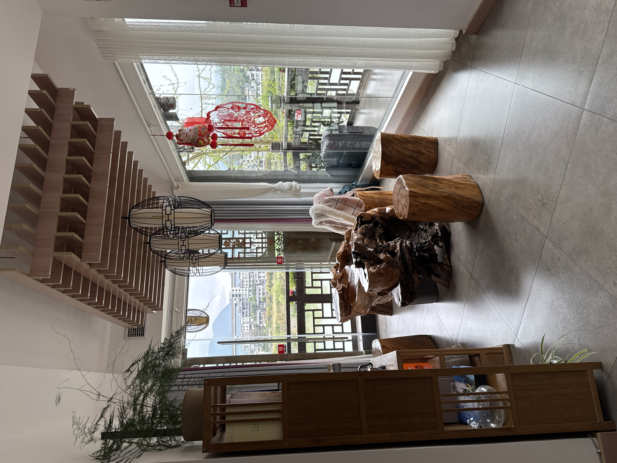 The image shows an interior scene, likely a living room, in Zhuoshui, China. The room features a decorative wooden ceiling, large windows offering a view of the town and distant mountains, log stools, and a large gnarled wooden table. A red papercut decoration hangs near the window. The floor is tiled. A wooden shelving unit holds books and decorative items. A potted plant is visible near the shelves. A suitcase is visible outside on the balcony. No people are visible.