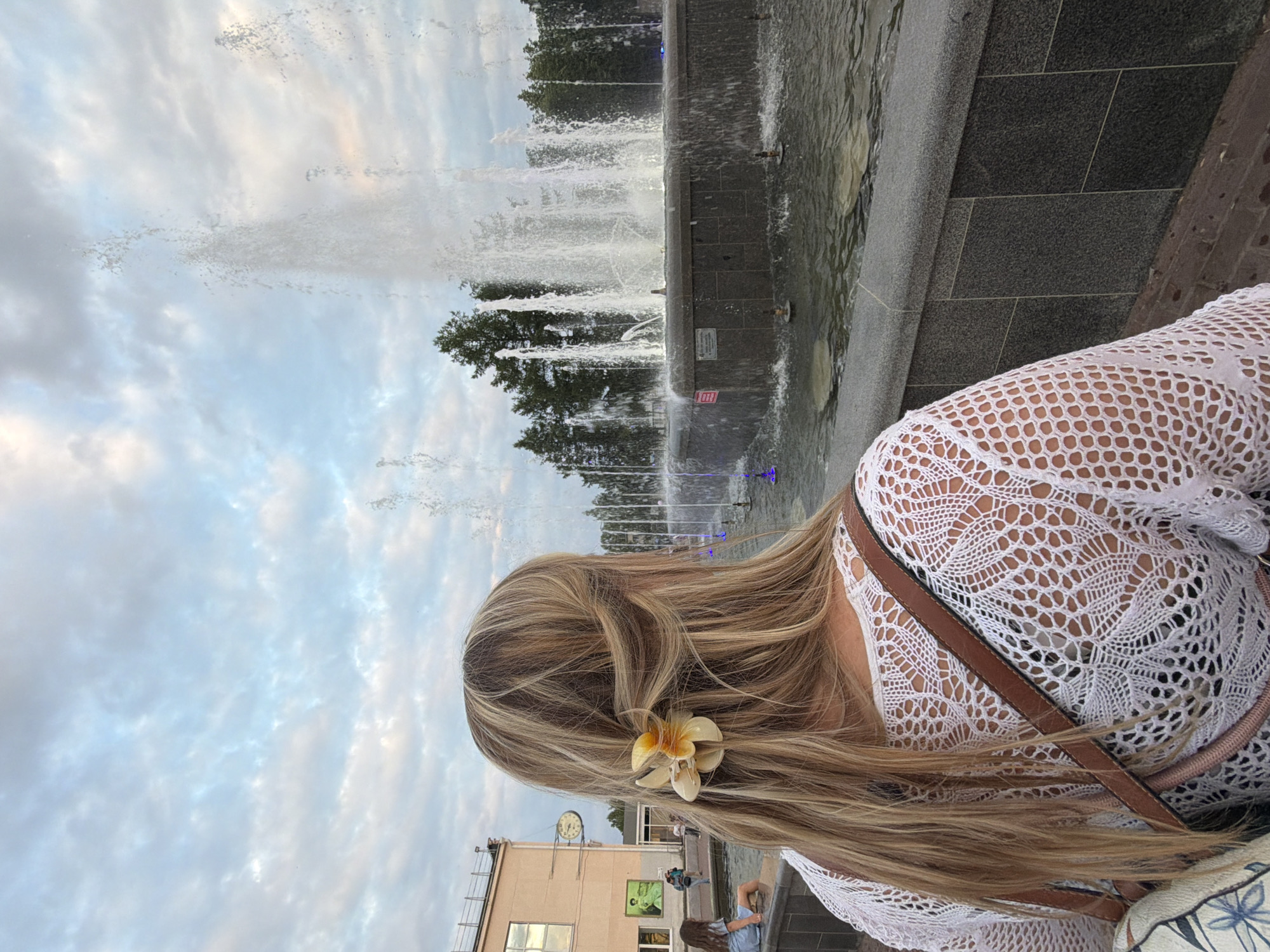 A young woman with blonde hair, adorned with a yellow and white flower clip, faces away from the camera. She is wearing a white, intricately crocheted top and a brown shoulder bag. She stands near a large, multi-tiered fountain, which is actively spraying water into the air. The fountain is surrounded by dark grey stone and a body of water. In the background, trees line the perimeter of the fountain area, and beyond that, a light-colored building with windows is visible, along with a clock face on its facade. The sky above is filled with clouds, suggesting it is either early morning or late afternoon. The scene is set in the city of Chaikovsky, Russia, likely in a public square or park.