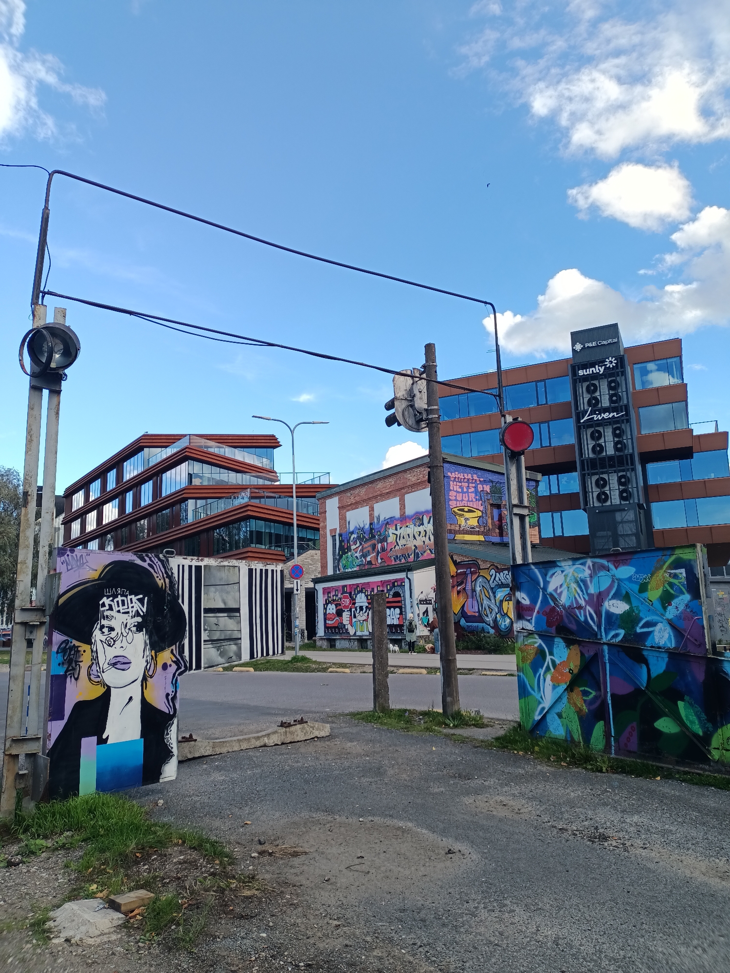 The image depicts an urban outdoor scene in Tallinn, Estonia, likely in a modern business district or a renovated industrial area, given the presence of contemporary architecture alongside graffiti art. In the foreground, a traffic light with a red signal is visible, mounted on a pole. To the left of the traffic light, a large, stylized graffiti mural of a woman's face wearing a wide-brimmed hat is painted on a surface that appears to be a shipping container or a similar structure. Further to the left, a vertical pole with an old-fashioned traffic signal head is present.
Behind the foreground elements, several buildings are visible. The most prominent is a multi-story building with a modern design, featuring a facade of brown wood-like material and large, reflective glass windows. On this building, prominent signage includes "P&E Capital" at the top and "sunly liven" below, suggesting it is an office building or a commercial complex. To the left of this building, another modern building with similar glass-paneled architecture is partially visible.
Interspersed between the modern buildings and the foreground graffiti are more examples of street art. A brick building in the middle ground is covered in colorful graffiti, and a separate, blue-painted structure on the right is also adorned with vibrant floral graffiti. The ground is paved with asphalt and gravel. The sky above is blue with scattered white clouds, indicating fair weather. The overall impression is one of a dynamic urban environment that blends contemporary development with vibrant street art culture.