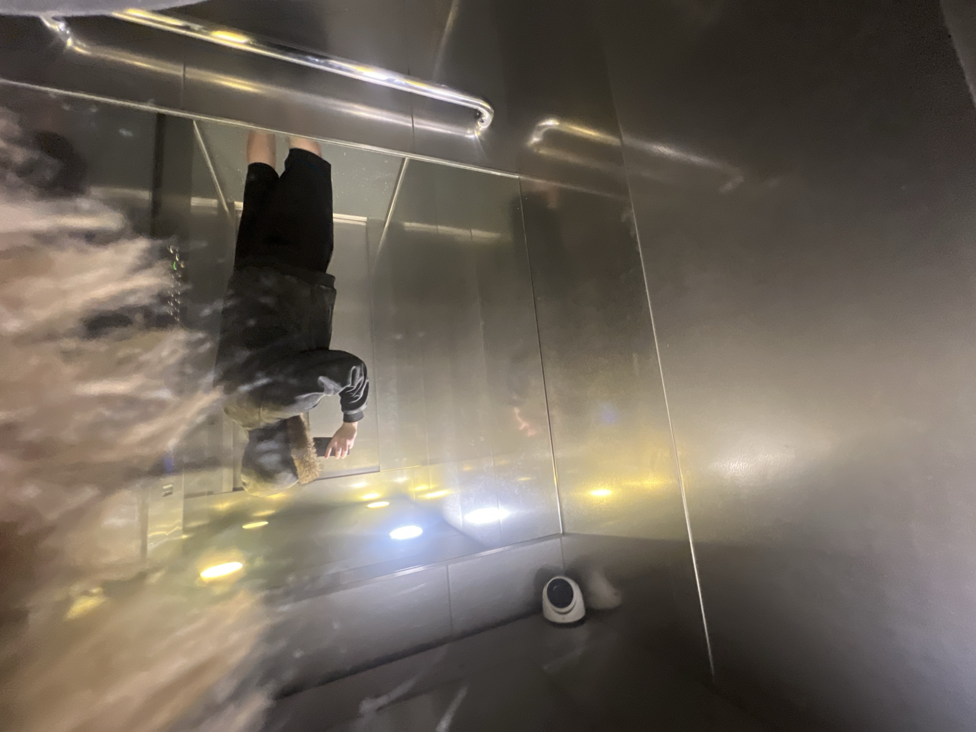 The image displays the interior of an elevator, rotated 90 degrees counter-clockwise from its upright orientation. The elevator features highly reflective, metallic walls, creating strong reflections of its contents and lighting.
Prominently reflected on the right side of the image is an upside-down person. This individual is dressed in dark clothing, including a long-sleeved top and what appears to be a fur-lined hood or hat. They are holding a mobile phone in their right hand, suggesting they might be viewing the screen or taking a picture.
A shiny, metallic handrail runs horizontally across the upper-middle section of the frame. Multiple bright, circular and oval lights, presumably from the elevator's ceiling, are reflected on the lower part of the reflective wall, casting luminous spots. A small, white security camera is visible on the floor in the bottom-right corner of the image.
A significant portion of the left side of the image is covered by a soft, light-colored blur or smudge, creating a diffused, almost ethereal effect that partially obscures the reflections in that area. No discernible text is visible anywhere in the frame. The overall setting is an artificially lit indoor environment, consistent with an elevator in an urban location such as Saint Petersburg, Russia.