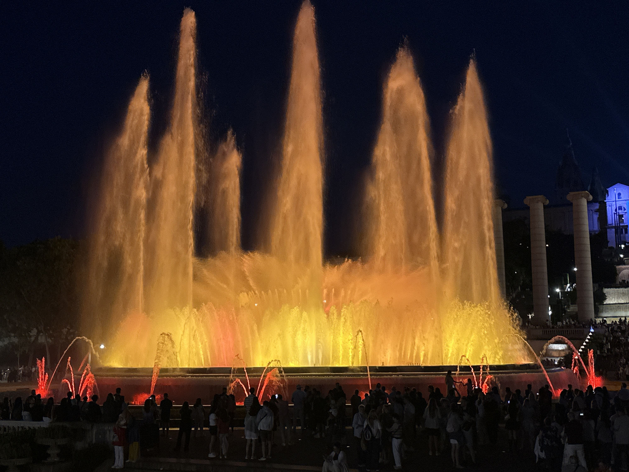 This image captures the illuminated Magic Fountain of Montjuïc in Barcelona, Spain, at night. The central focus is a large water fountain display with numerous jets of water. Taller, vertical plumes of water in the background are brightly lit in shades of orange and yellow, creating a radiant glow against the dark night sky. In the foreground, smaller, arching water jets are intensely illuminated in red.
A substantial crowd of people is gathered around the perimeter of the fountain, predominantly in the lower half of the image. They are standing, facing the fountain, observing the water and light show. Their forms are largely silhouetted or dimly lit due to the night setting, with few discernible individual details or specific actions.
In the upper right background, architectural structures are visible, including several large, pale pillars and a building with blue-tinted lighting further in the distance. The sky above is a deep, dark blue, indicating nighttime. The overall scene is a vibrant public spectacle, with the illuminated fountain dominating the composition and attracting a large audience.