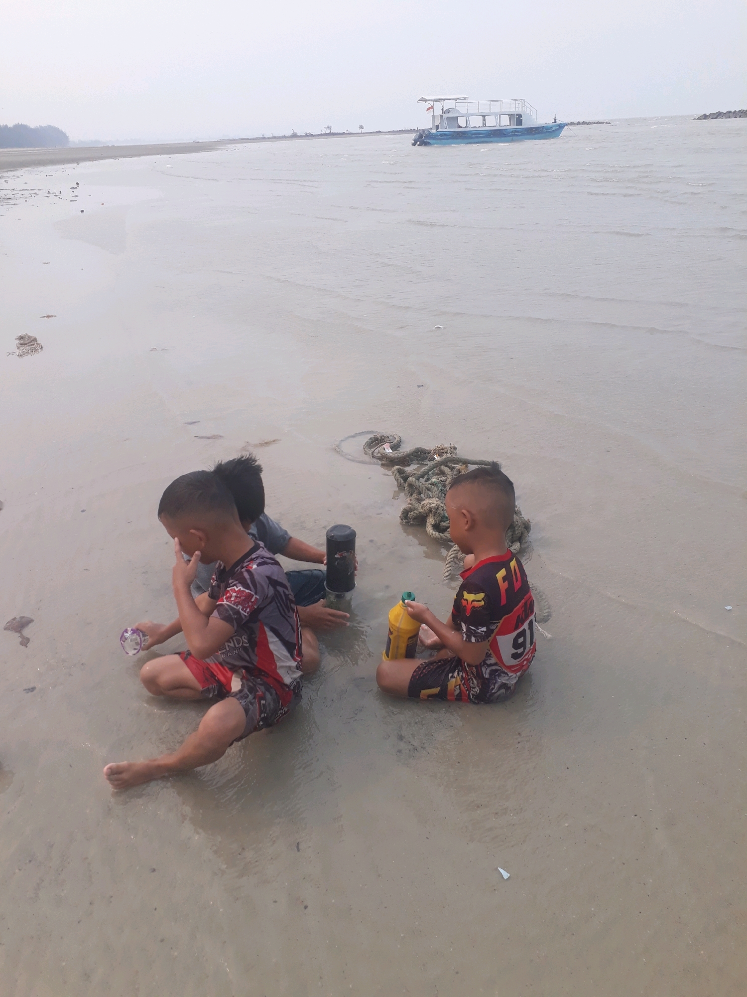 The image depicts a serene beach scene at what appears to be low tide, with shallow water lapping at the sandy shore. In the foreground, three young boys are sitting on the wet sand, engaged in play. The boy on the left, wearing a red, white, and black patterned t-shirt and shorts, is holding a small, clear plastic container and appears to be looking down at it. The boy in the middle, wearing a gray t-shirt, is partially obscured by the boy to his left, and is reaching towards a black cylindrical object. The boy on the right, dressed in a red and black shirt with the number "91" prominently displayed and matching shorts, is holding a yellow bottle and looking towards the water. Scattered around them are a few pieces of debris, including a coiled rope and some small natural items. In the middle ground, a blue and white boat with a canopy is anchored offshore, and breakwaters are visible on either side of it, suggesting a somewhat protected area. The background reveals a hazy, overcast sky and a distant shoreline with a line of trees. The overall impression is one of a calm, everyday moment captured at a coastal location, likely within or near the city of Pangkalpinang, Indonesia, given the presence of such a boat and the typical coastal environment.