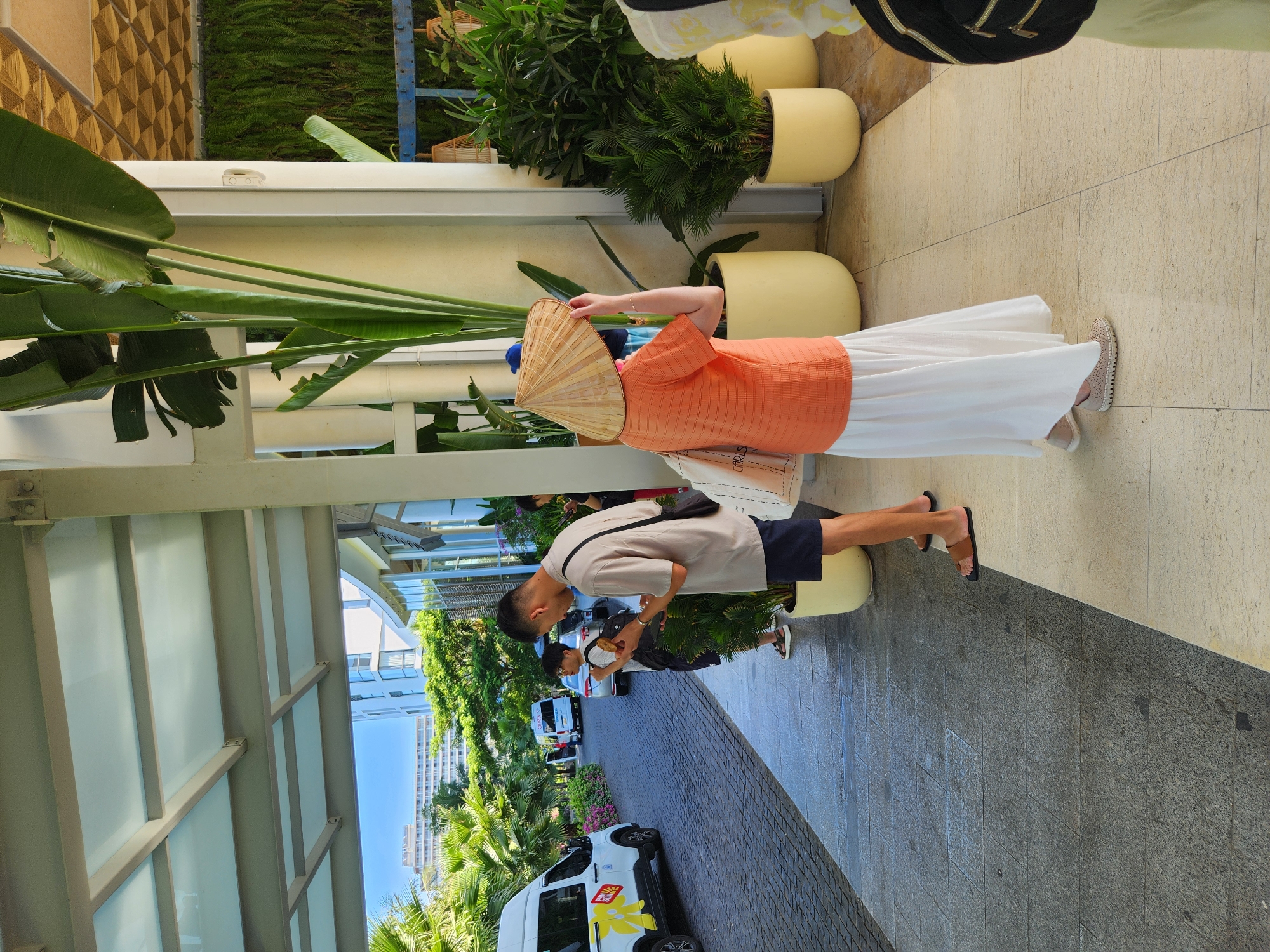 In Đà Nẵng, Vietnam, an outdoor scene unfolds under a covered walkway of a building, likely a resort or hotel entrance. Sunlight casts shadows across the tiled walkway. Two men and a woman are visible. One man, wearing a beige t-shirt and dark shorts, stands looking at his phone while holding a snack. He wears brown sandals. Beside him, a younger man with glasses, wearing a black t-shirt and shorts, holds a bag. He also wears sandals. Further down the walkway, a woman in an orange shirt and a long white skirt, wearing a conical hat, stands with her back to the camera, holding a beige canvas bag. In the background, a white van with "FUNNY SUN" branding is parked on the paved road. Lush green palm trees and foliage surround the area, with a building visible in the distance. Large potted plants, including a tall palm-like plant, are placed near the entrance. The overall atmosphere suggests a warm, tropical environment.