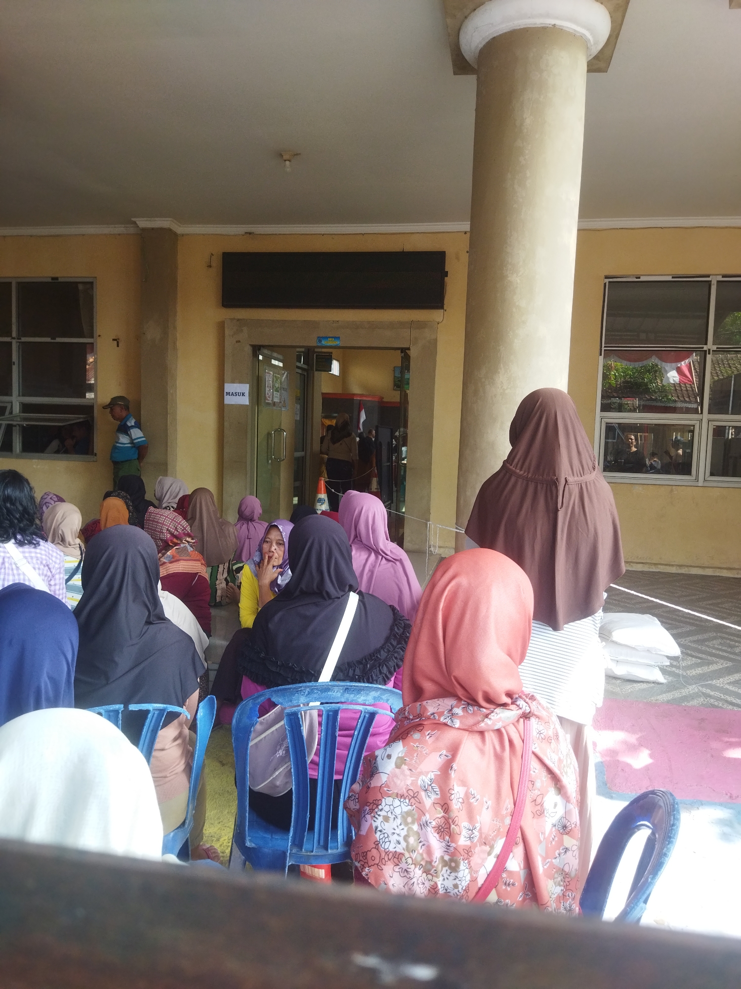 The image depicts an outdoor waiting area, possibly at a government office or public service facility, in Banjar, Indonesia. A group of people, predominantly women wearing headscarves and modest clothing, are seated and standing, awaiting services. The scene is characterized by a large, light-colored pillar dominating the center-right of the frame, suggesting an architectural element of the building. Yellow walls surround the area, with windows reflecting outdoor elements, including trees and structures. A sign reading "MASUK" (meaning "Entrance" in Indonesian) is visible near a doorway, indicating an entry point. Some individuals are looking towards the entrance or conversing, while others appear to be simply waiting. The ground is paved, with some areas of colorful markings, possibly for queuing or designated waiting spots. Bags and chairs, some of which are blue plastic, are present in the scene. The overall atmosphere suggests a mundane, everyday activity of people waiting for a public service.
