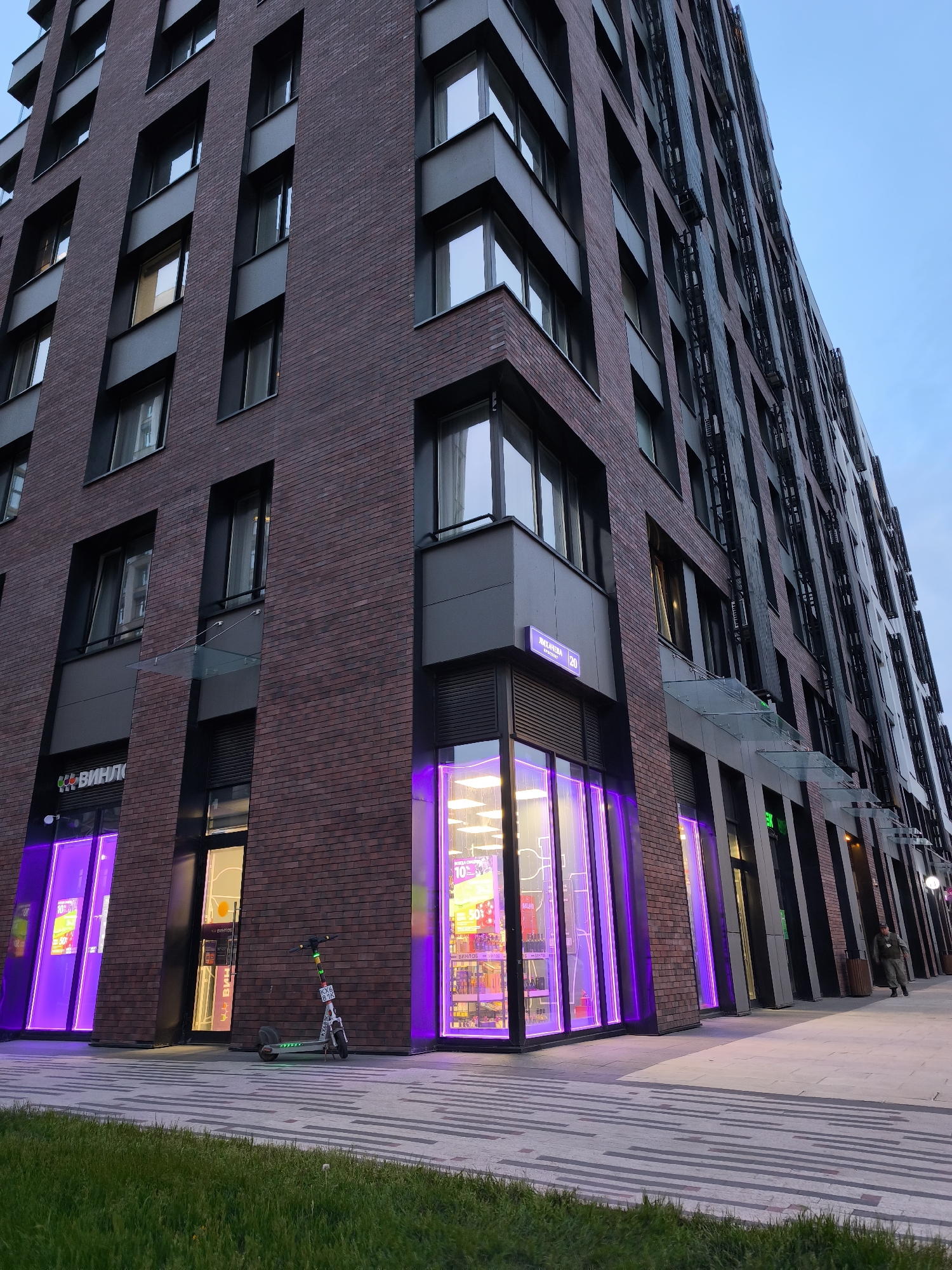 The image shows the exterior of a modern, multi-story building, likely in a commercial area. The building has a brick facade and dark window frames. The street-level storefronts are illuminated with purple neon lights, drawing attention to the businesses inside. A street sign is visible on the building. An electric scooter is parked nearby. A person walks near the building. The street and sidewalk are paved with a patterned design, and a patch of green grass is visible at the bottom left. The sky is a pale blue. The location is Moscow, Russia as is indicated by the street sign with Russian lettering on it.