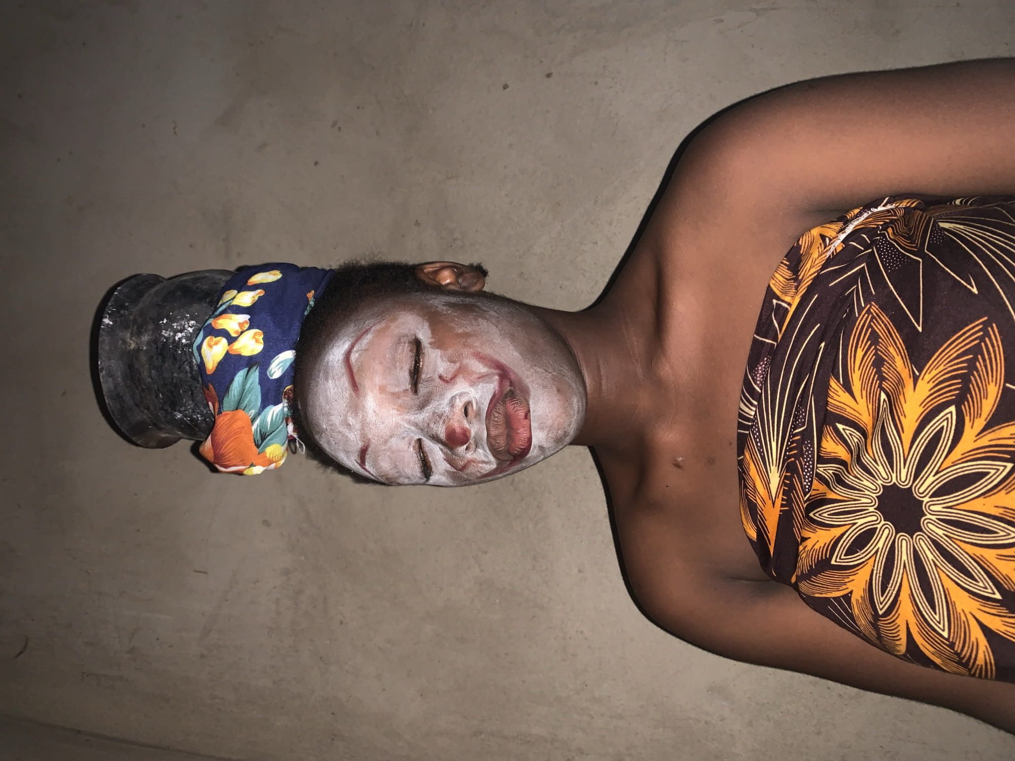 An overhead shot shows a woman lying supine on a beige, textured surface. The woman is dark-skinned and wears a white facial mask, which covers most of her face and lips, leaving some areas of skin exposed. She also wears a blue headscarf with orange and yellow floral patterns. Her shoulders are bare, and an orange and black floral patterned cloth is wrapped around her upper body. To the left of her head, there is a black object that may be a mortar and pestle.
The location is Agban, Nigeria.