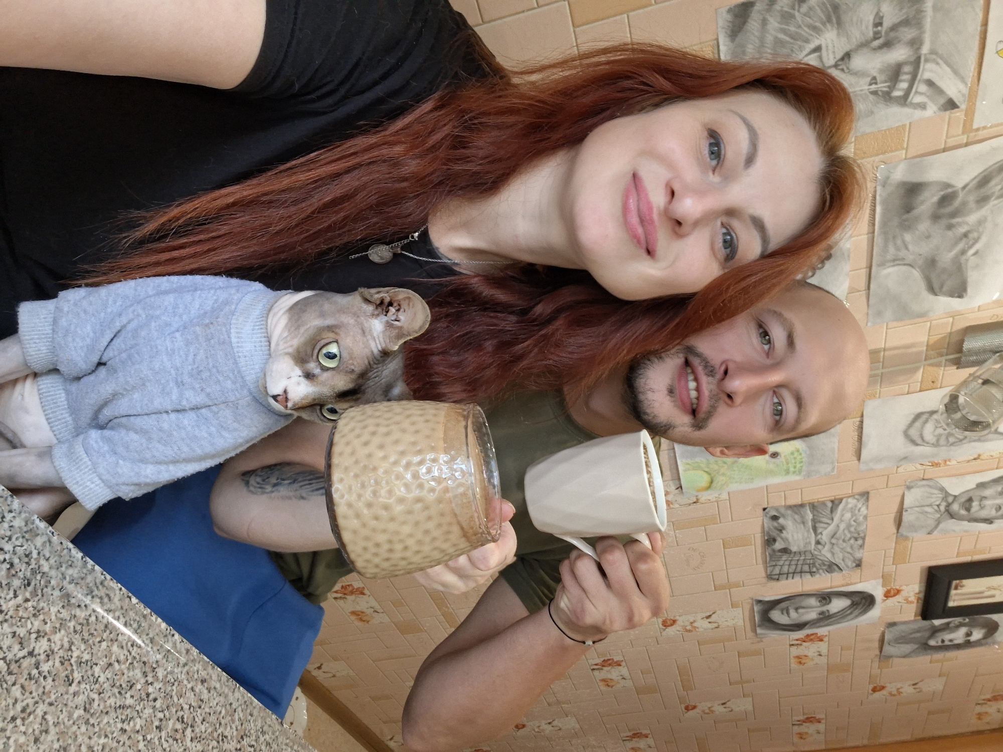 In a room with patterned wallpaper, a man and a woman are taking a selfie. The woman, on the left, has long, reddish-brown hair and is wearing a black t-shirt. She is smiling at the camera. The man, on the right, is bald and has a mustache. He is wearing a dark green t-shirt and is holding a white mug of coffee. Between the couple, a Sphynx cat wearing a gray sweater is visible. The cat is looking towards the right. On the wall behind them, several charcoal drawings are hanging. The foreground shows a speckled countertop. The scene appears to be indoors, possibly in a kitchen or living area. There is no information within the image to suggest the location is in Myadzyel, Belarus.
